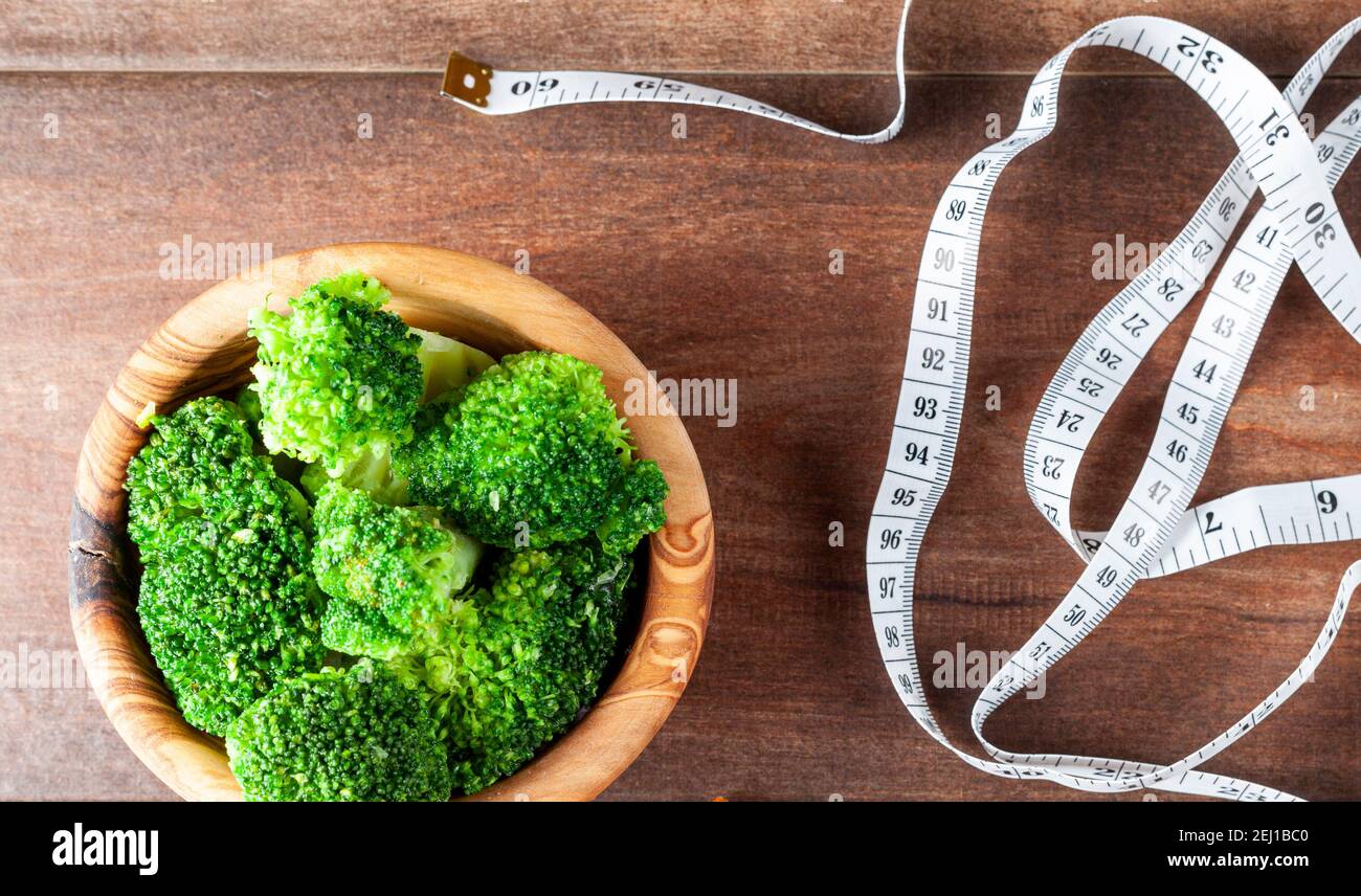Immagine di una ciotola di legno di fiori freschi di broccoli e un nastro di misurazione fianco a fianco sulla superficie di legno. Immagine concettuale per dieta, fitness, noi Foto Stock