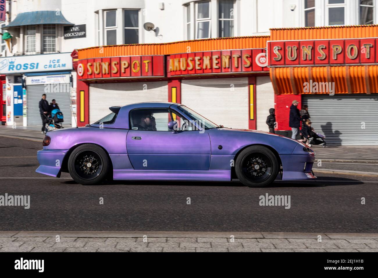 Auto Mazda MX5 personalizzata che passa accanto a portici chiusi a Southend on Sea, Essex, Regno Unito. Sfumature di viola vernice personalizzata con finitura ruvida. Giorno di sole Foto Stock