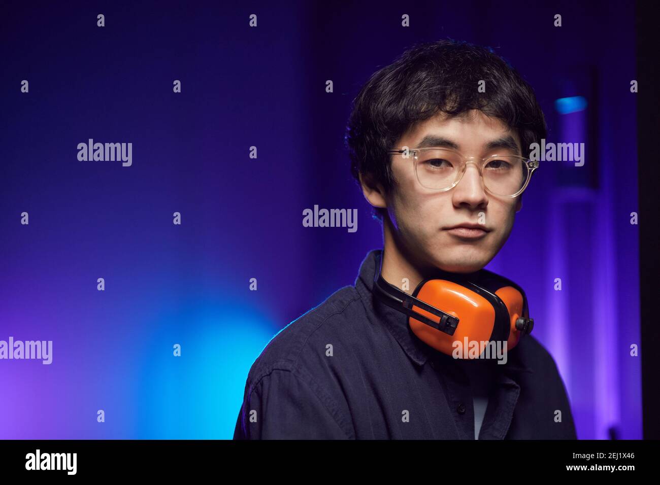 Ritratto di un giovane asiatico che indossa le cuffie guardando la fotocamera mentre si trova in un futuristico interno illuminato da luce blu, spazio copia Foto Stock