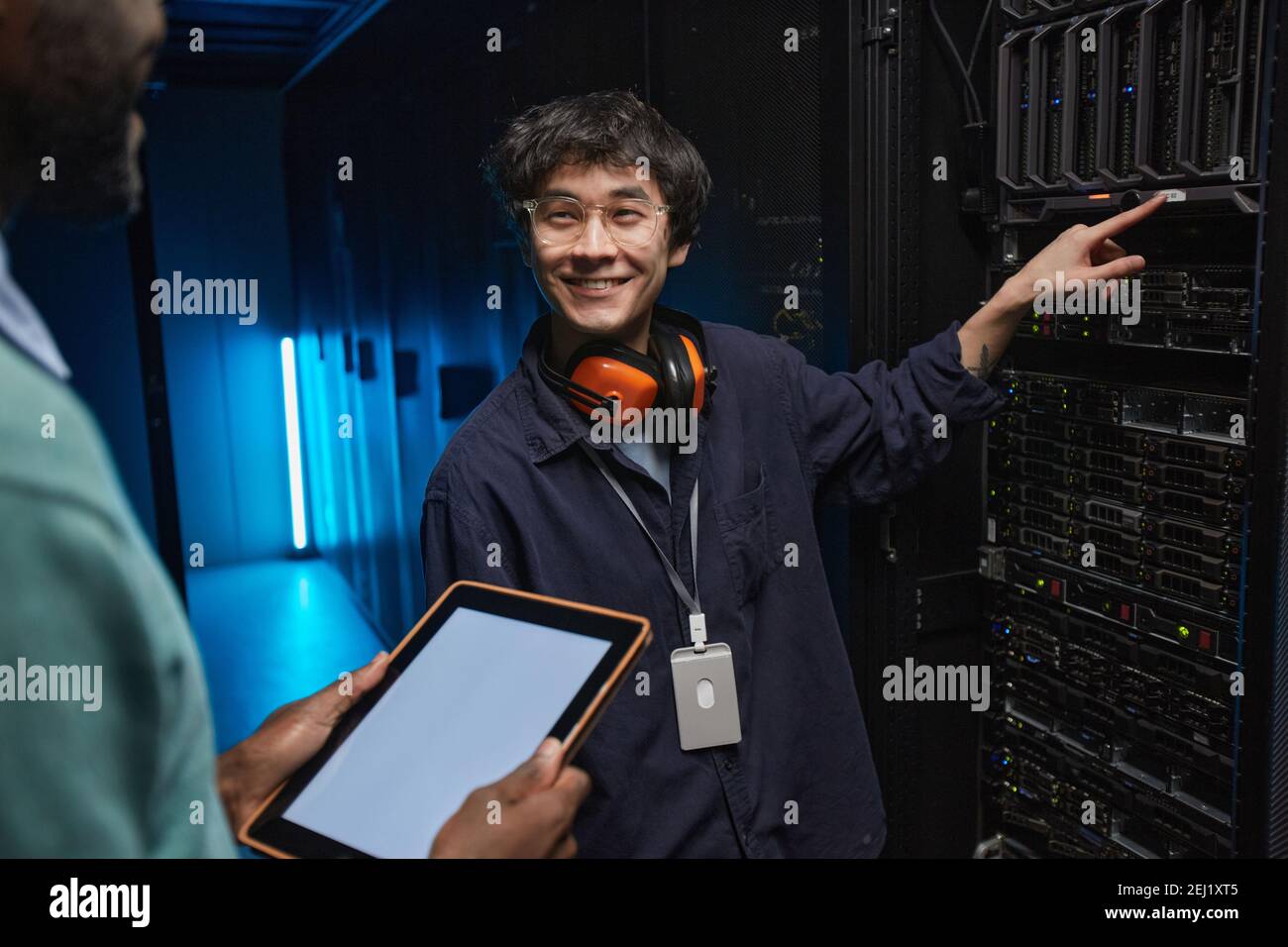 Ritratto di un uomo asiatico sorridente che guarda un collega durante la configurazione di server di supercomputer nel data center, spazio di copia Foto Stock