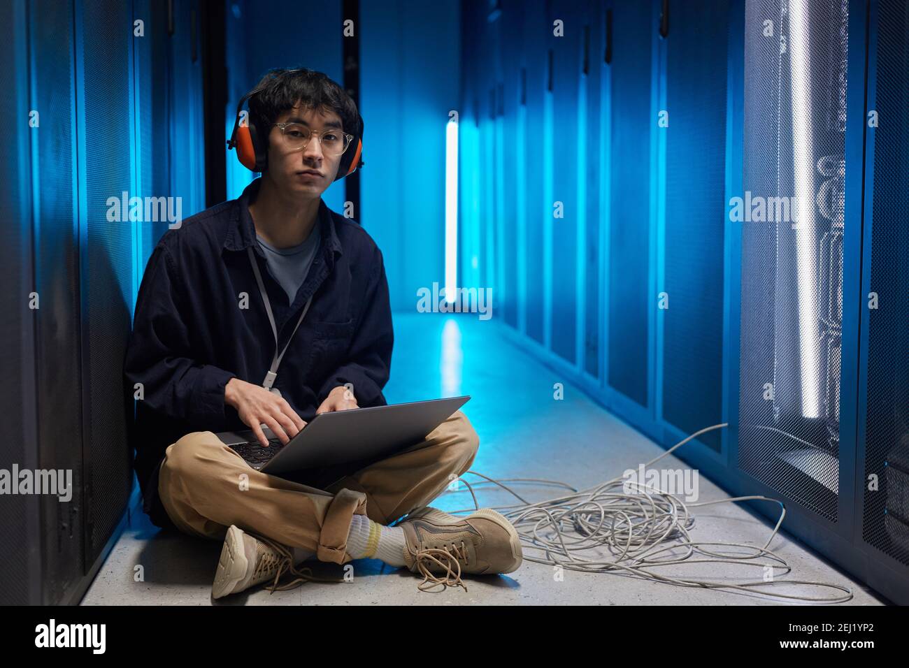 Ritratto a lunghezza intera di giovane asiatico seduto sul pavimento nella stanza del server illuminato da luce blu durante la configurazione della rete di supercomputer, spazio di copia Foto Stock