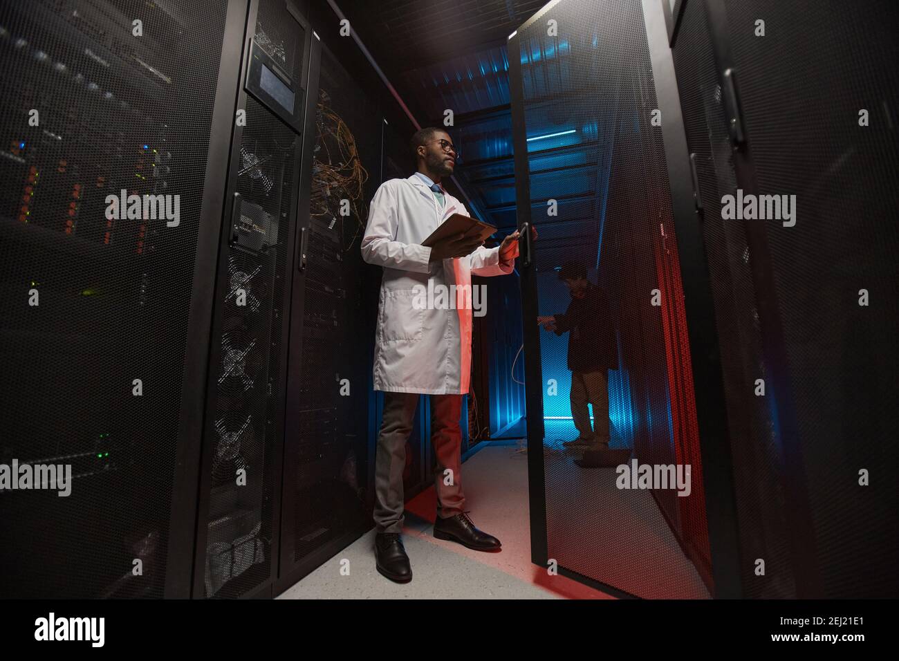 Ritratto a tutta lunghezza di uno scienziato di dati afro-americano indossando camice da laboratorio mentre si lavora con supercomputer in sala server, spazio di copia Foto Stock