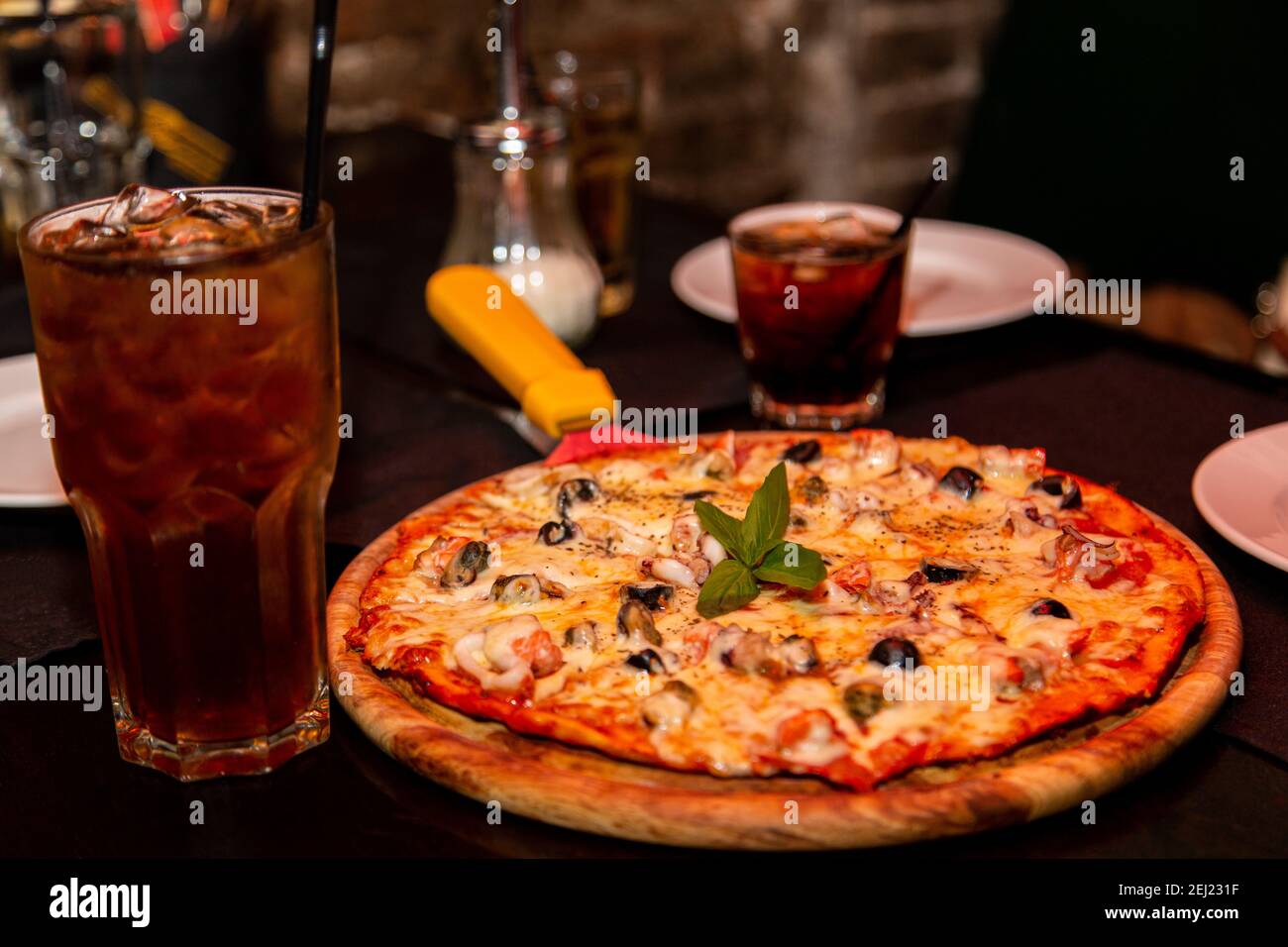Una pizza appetitosa con frutti di mare e formaggi si trova su un piatto di legno sul tavolo. Con una bevanda fredda marrone ghiacciata. Foto Stock
