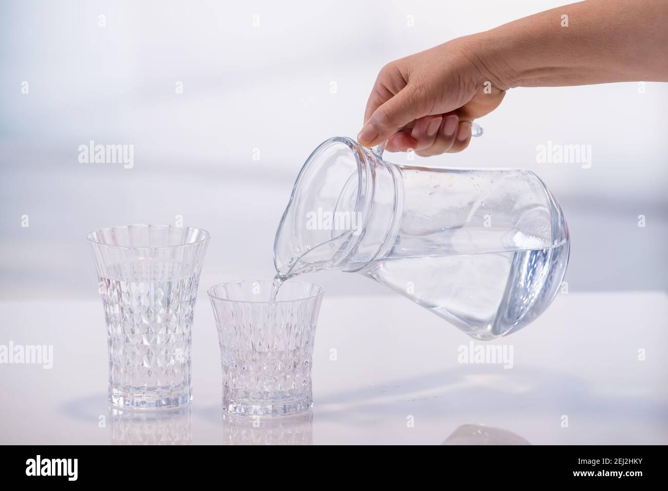 Mano femminile che tiene un vaso d'acqua e lo versa in un bicchiere su uno sfondo fuori fuoco. Messa a fuoco selettiva e primo piano. Concetto di acqua e salute. Foto Stock