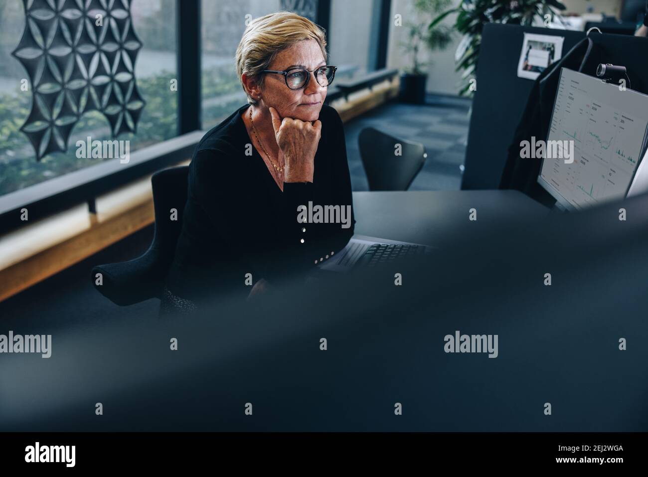 Donna seduta alla sua scrivania con la mano sul mento e che guarda il monitor del computer. donna d'affari seduta alla sua scrivania e a pensare. Foto Stock