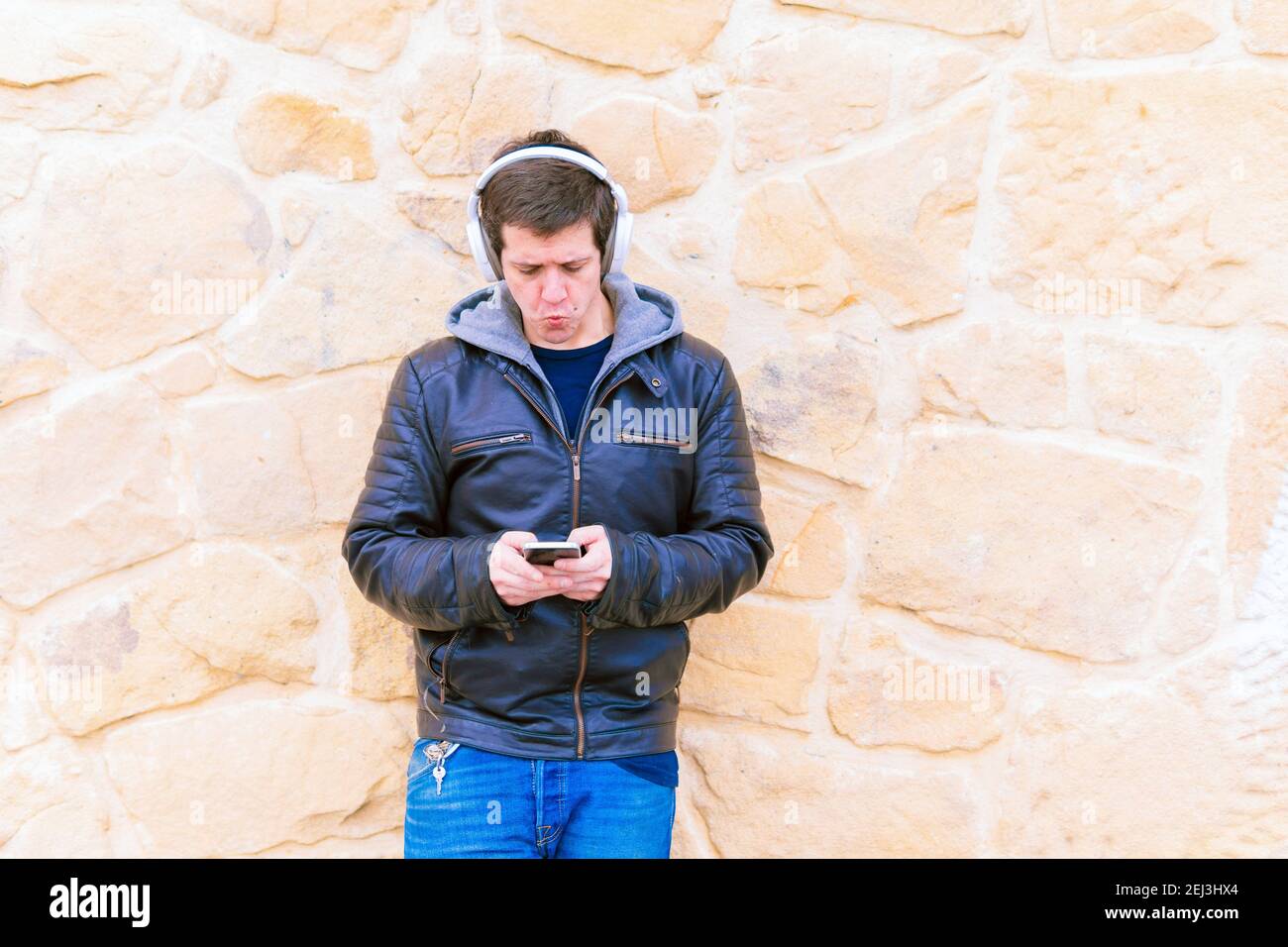 Attraente giovane uomo di mezza età appoggiato su una parete di pietra che indossa il wireless cuffie musicali e giacca in pelle con vista sullo smartphone Foto Stock