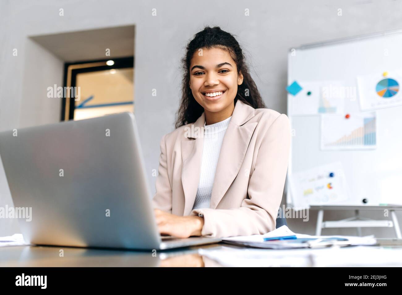 Elegante giovane donna d'affari si siede alla scrivania, sorridente e guarda la macchina fotografica. Una donna afro-americana che usa il laptop, sviluppa un nuovo progetto, navigando in internet Foto Stock