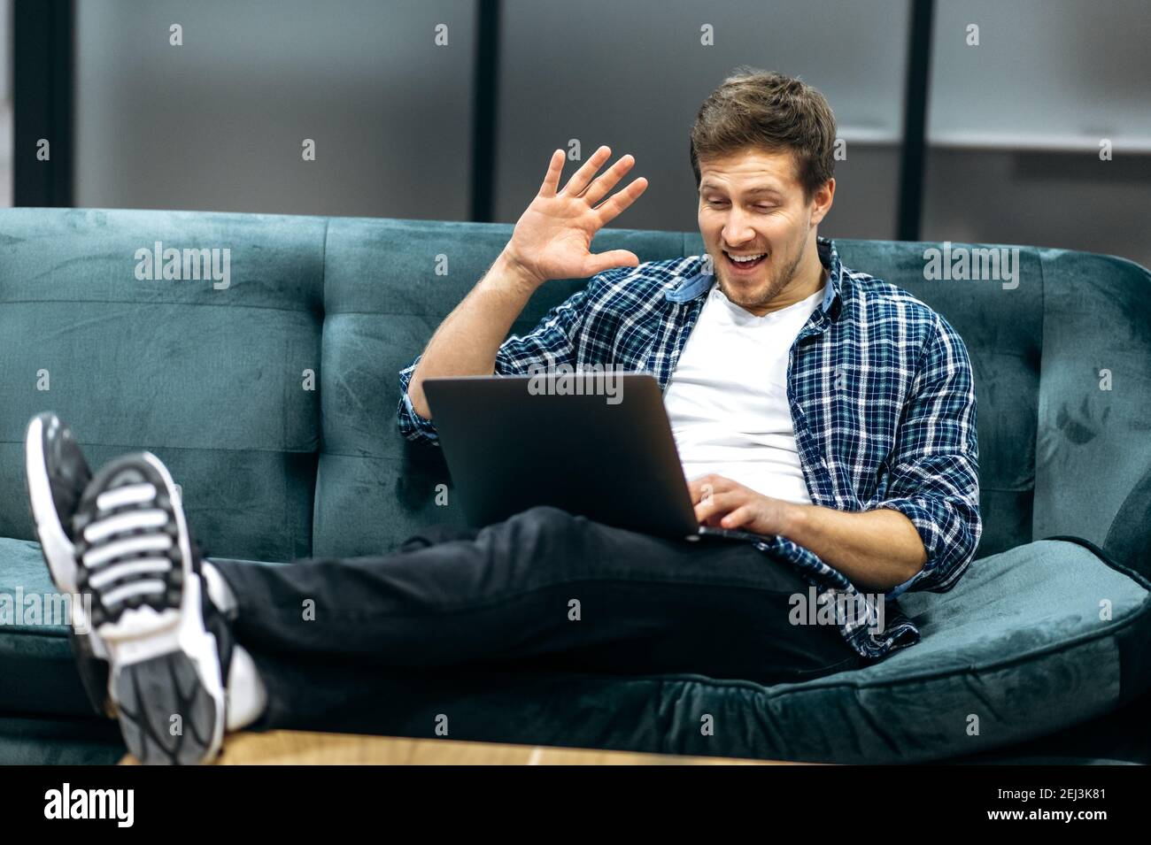 Gioioso freelance maschile o studente in un abbigliamento casual elegante seduto in un ufficio moderno, utilizzando un computer portatile. Il ragazzo comunica tramite videochiamata con colleghi o amici, sorridendo, concetto di comunicazione Foto Stock