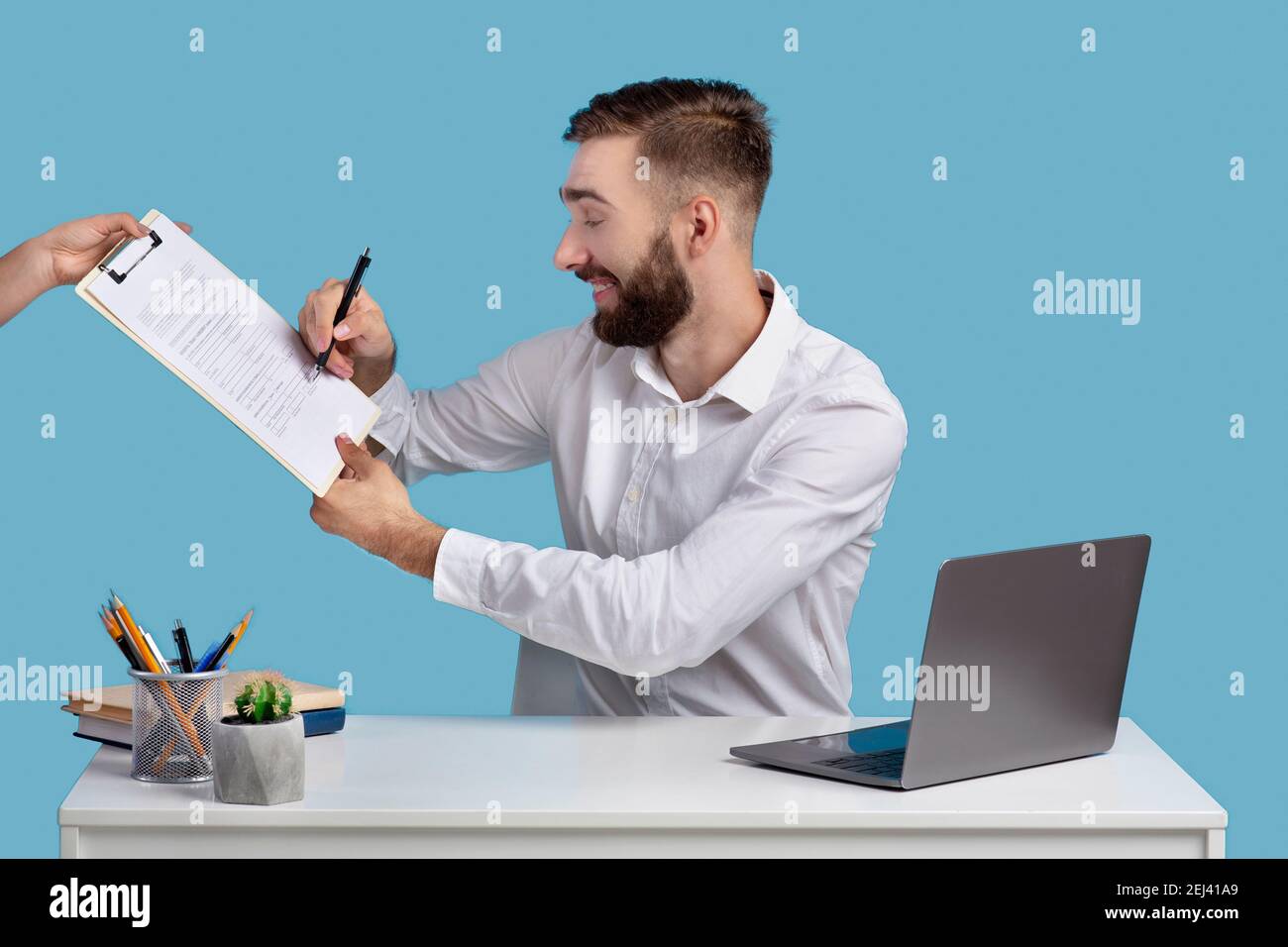 Uomo d'affari millenario in contratto di firma di abbigliamento formale o accordo alla scrivania in ufficio, sfondo blu studio Foto Stock