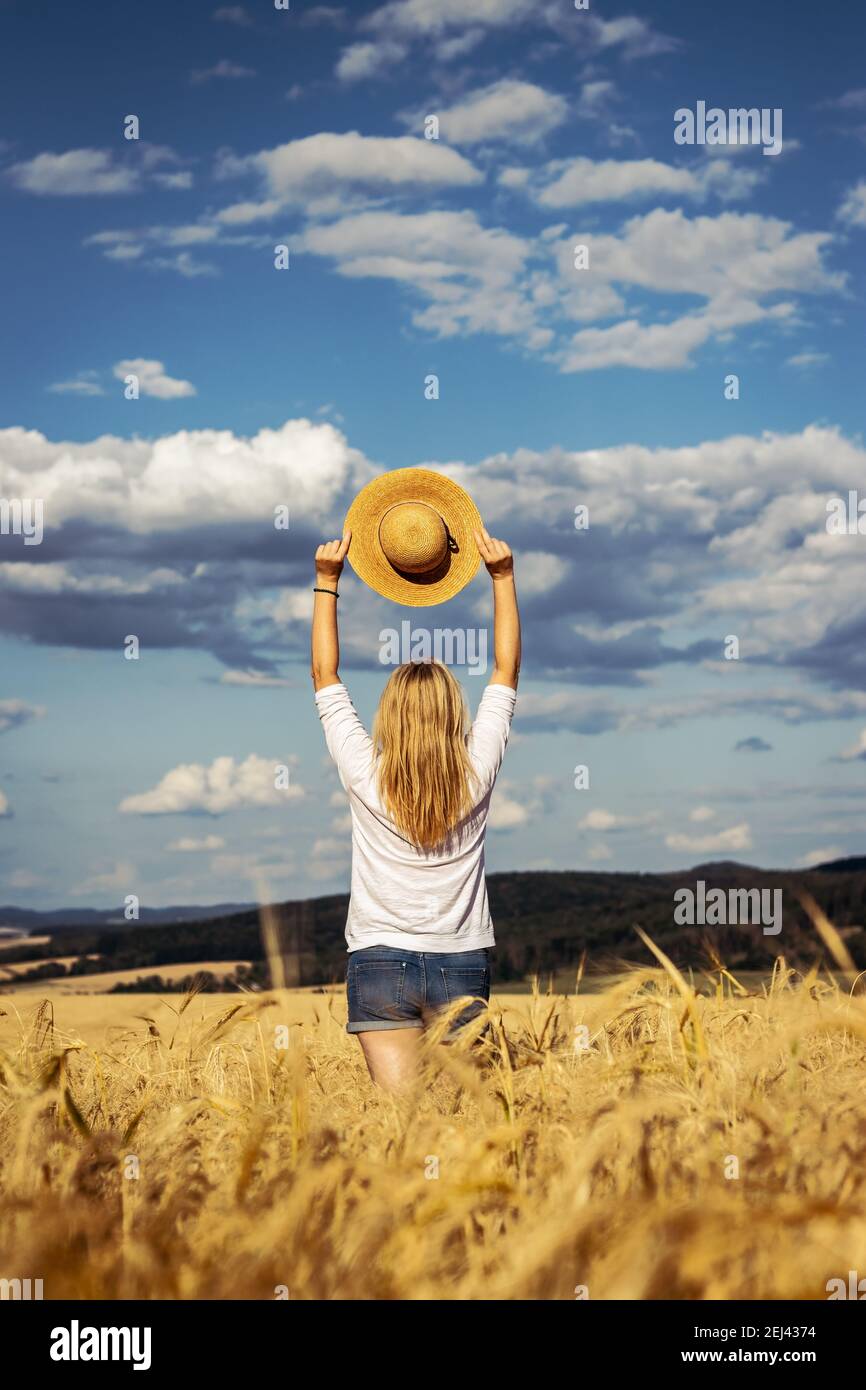 Donna felice che tiene cappello di paglia è in piedi in campo di grano. Godimento dell'estate. Donna con capelli biondi spensierati che indossa abiti casual contro il cielo bello Foto Stock