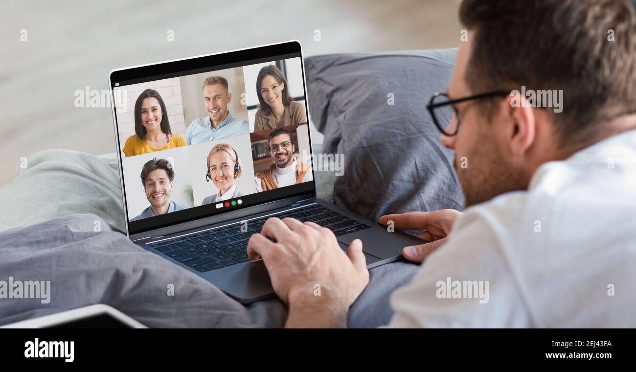 Giovane uomo che ha una riunione di lavoro on line su un computer portatile a casa, sdraiato a letto e che ha una videochiamata di gruppo con i suoi colleghi Foto Stock