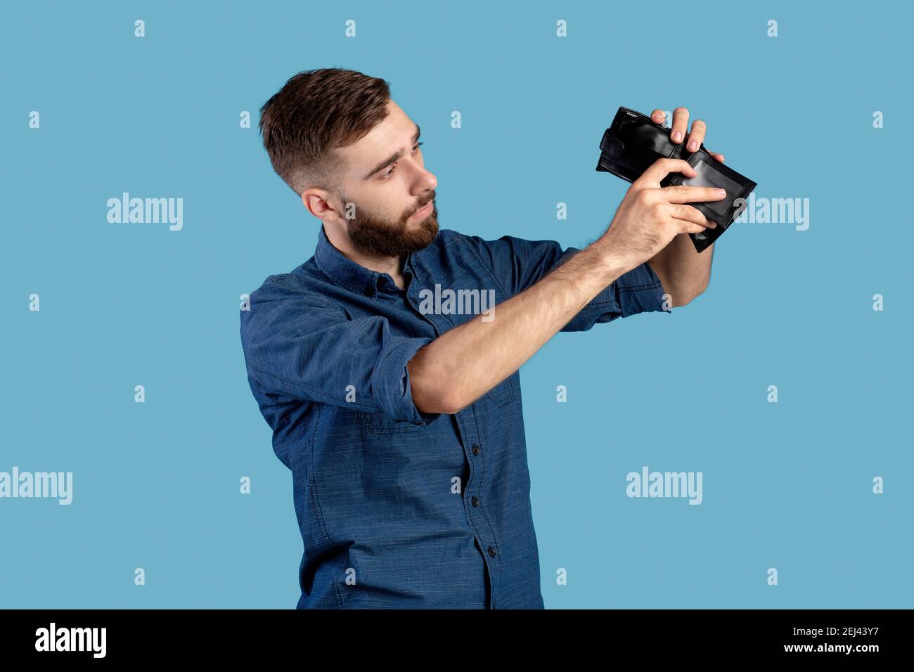 Fallimento, povertà e disoccupazione. Miserabile giovane uomo che guarda nel portafoglio vuoto senza soldi su sfondo blu Foto Stock