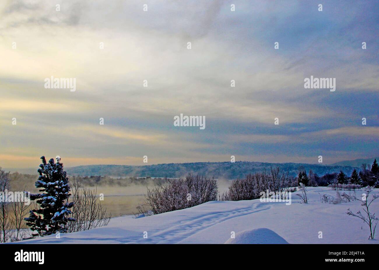 Vista su una riva del fiume innevata e montagne boscose al crepuscolo. Foto Stock