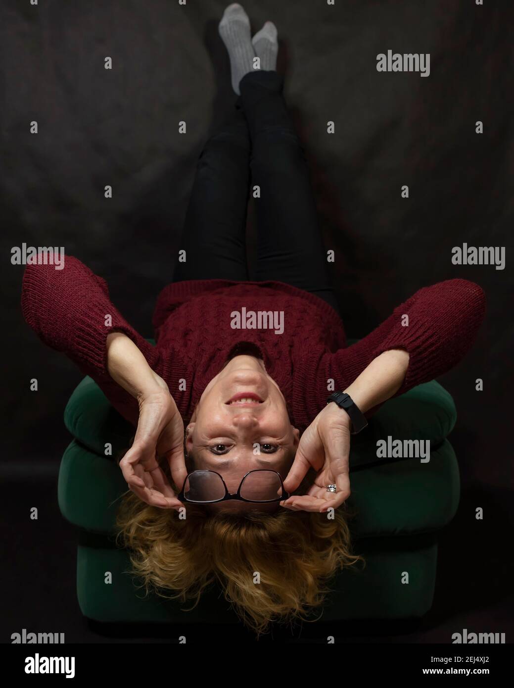 Una donna bianca di mezza età è sdraiata capovolta su un pouf mentre tiene i suoi bicchieri Foto Stock