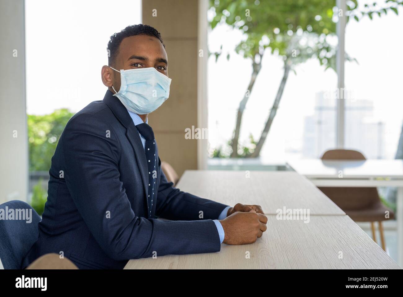 Uomo d'affari africano con maschera medica chirurgica per la protezione seduta a. ufficio Foto Stock