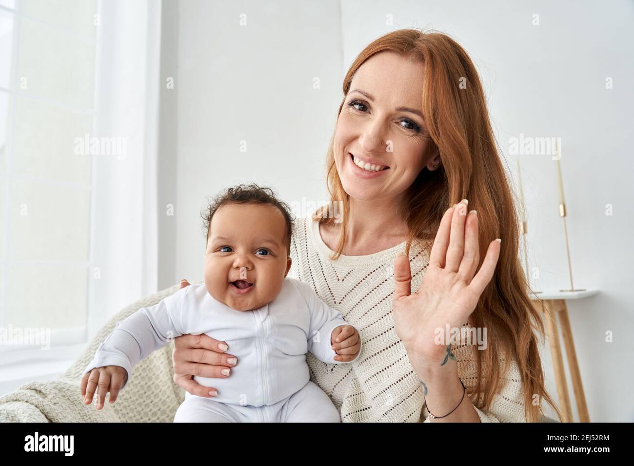 Madre felice che agita la mano che tiene la figlia del bambino divertente che guarda la macchina fotografica a casa. Foto Stock