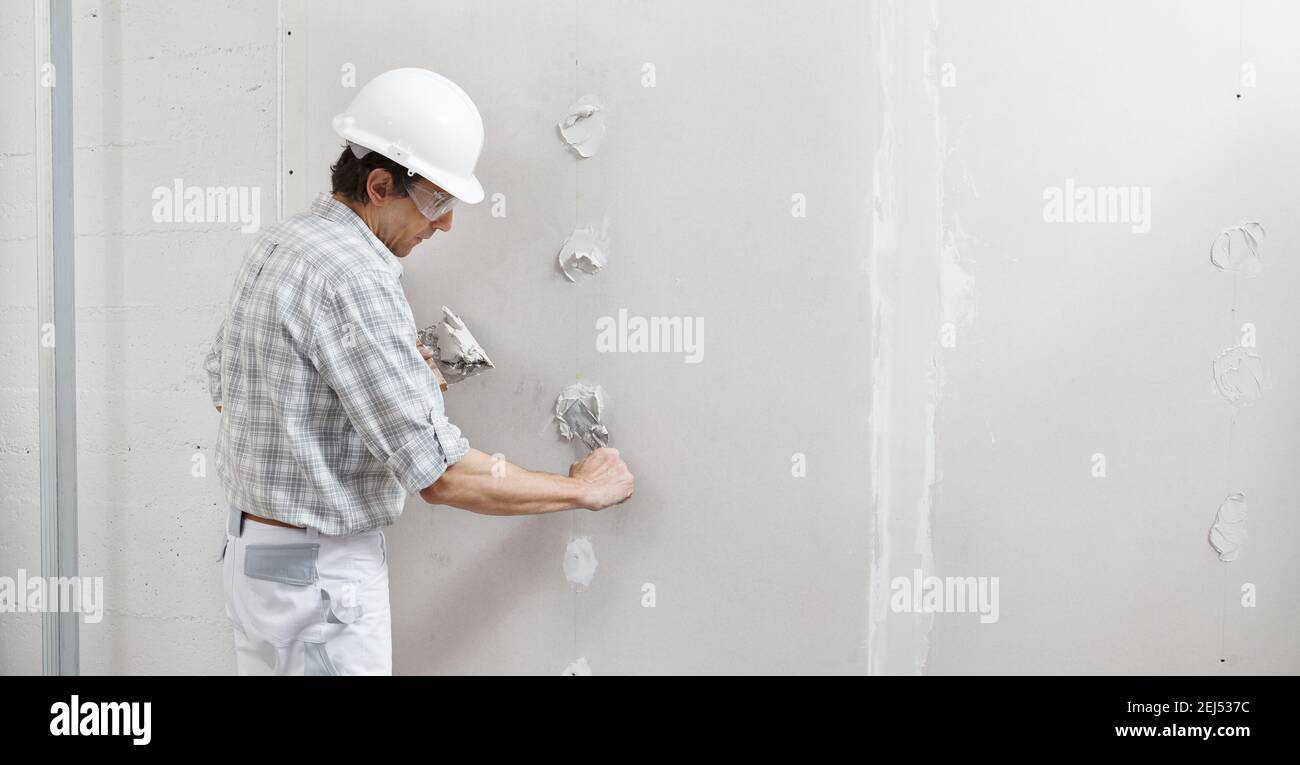 uomo di cartongesso operaio o intonaco mettendo intonaco su parete di cartongesso usando una cazzuola e una spatola, riempire i fori della vite, indossando hardhat bianco e saf Foto Stock