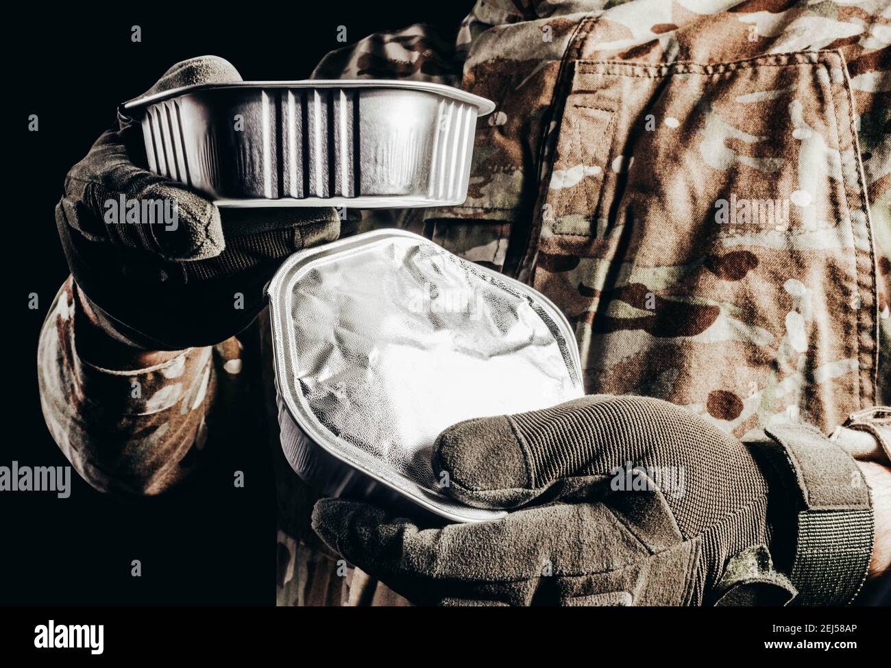 Foto del soldato in uniforme camuffata e guanti tattici che tengono in scatola il campo di cibo razione su sfondo nero. Foto Stock