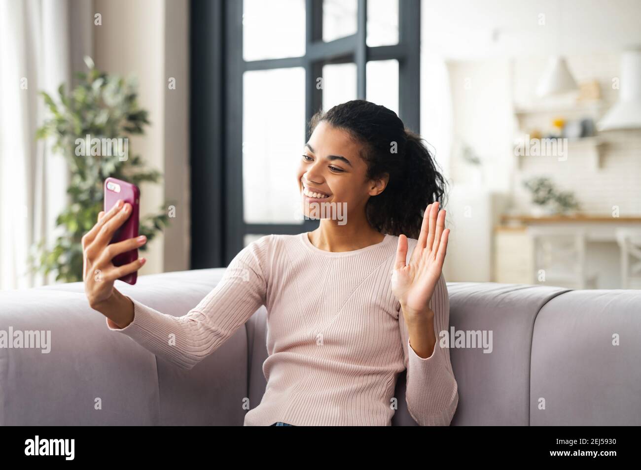 Giovane donna latina saluto da video link e sorridendo ampiamente, seduta su un comodo divano a casa, relazioni a lunga distanza, cucina leggera dietro Foto Stock