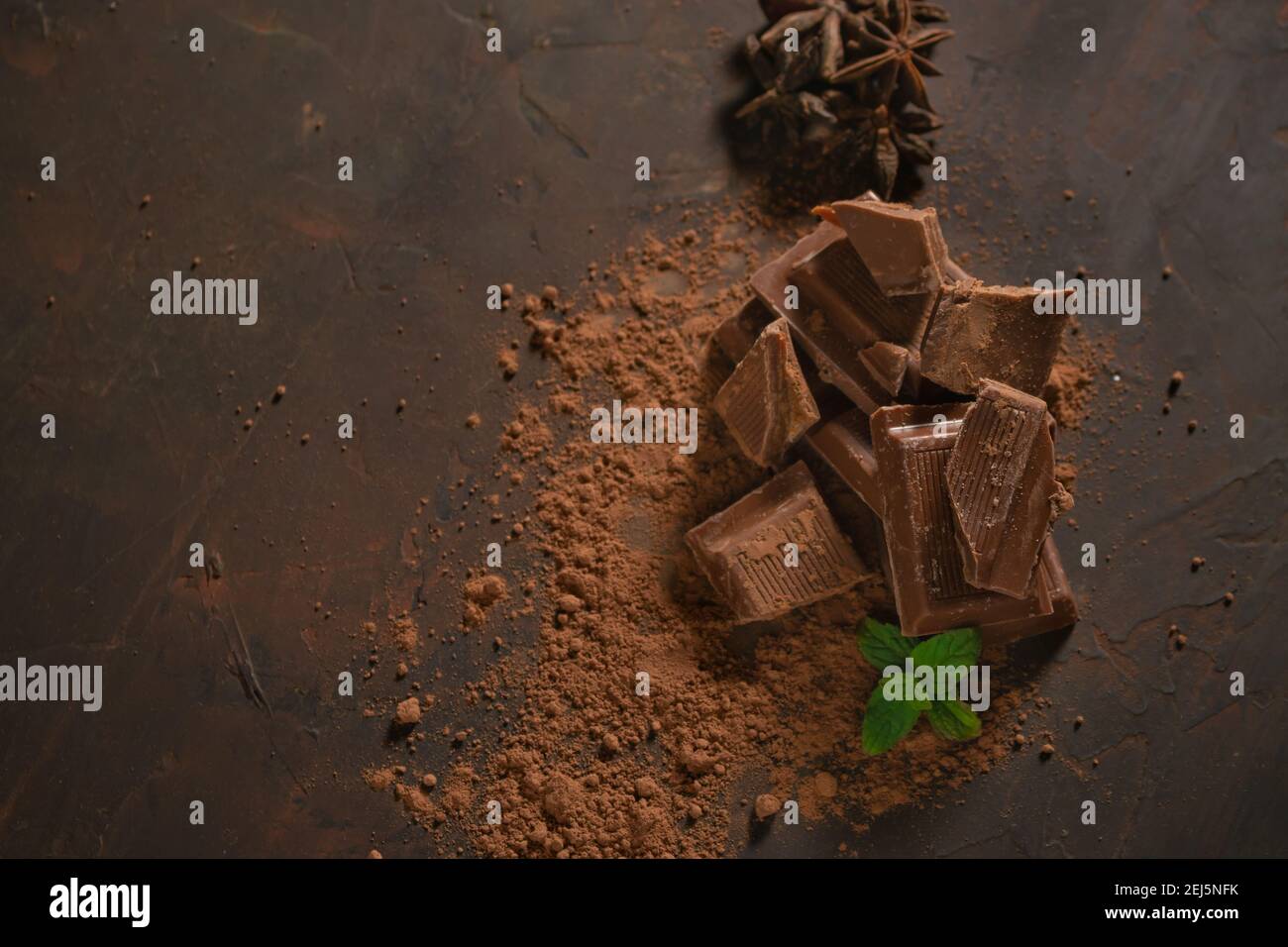 Diversi pezzi di cioccolato fondente e polvere di cacao su una superficie scura. Foglie di menta e semi di anice stellato completano l'immagine. Foto Stock