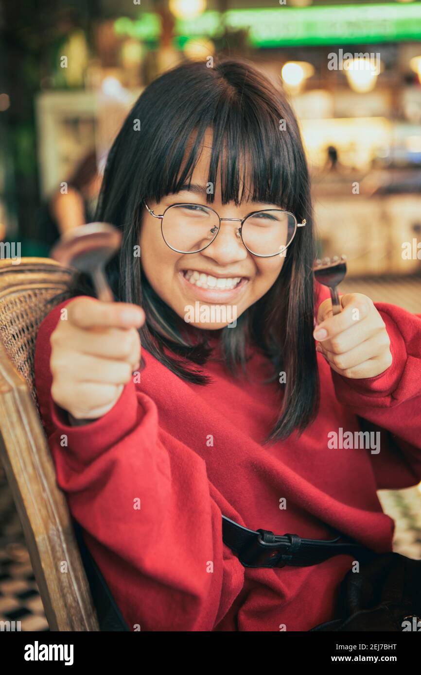 faccia divertente dell'adolescente asiatico solleva forchetta e cucchiaio sala da pranzo Foto Stock