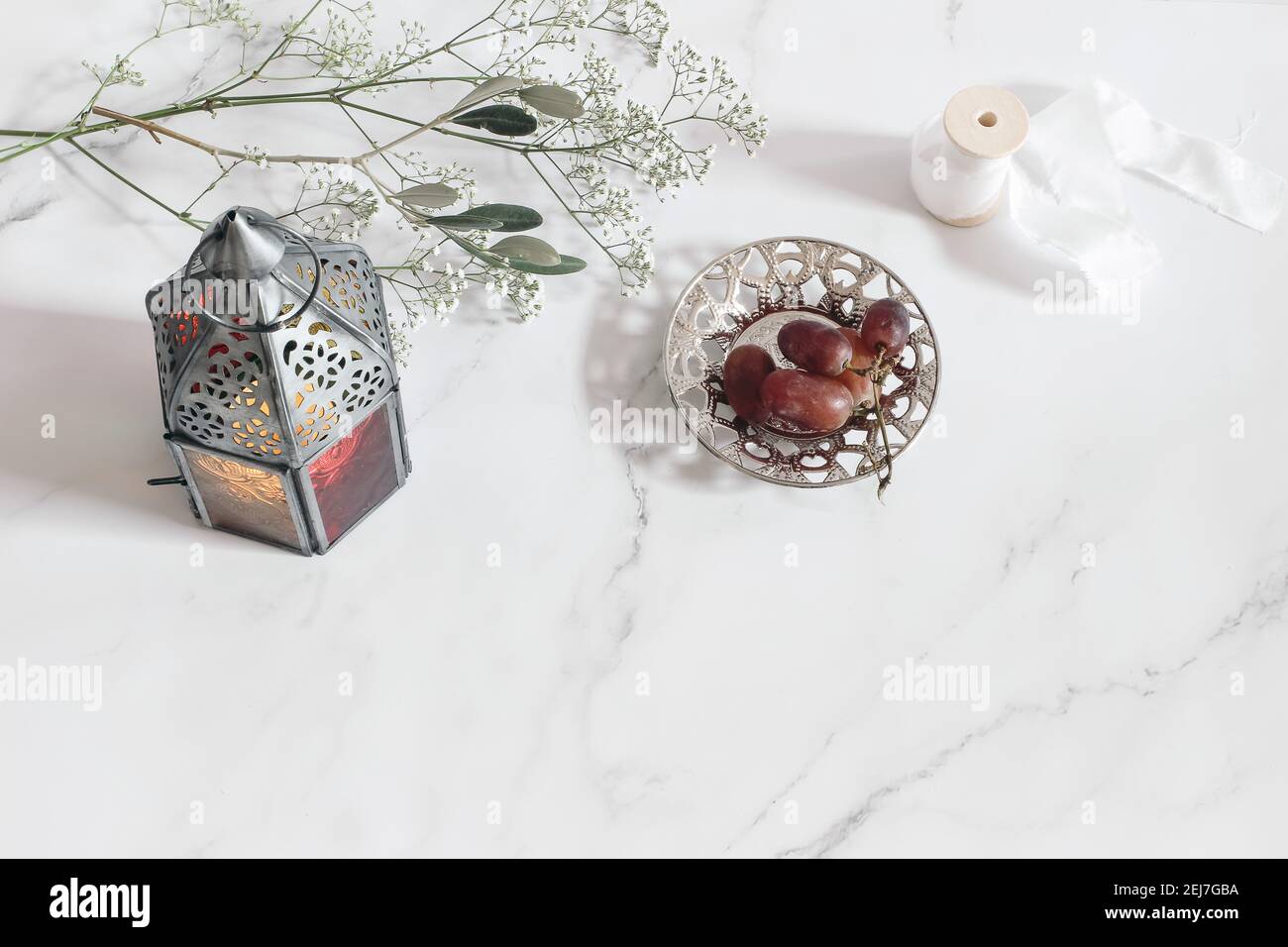 Marocchino d'epoca, lanterna araba. Ramo di ulivo, fiori di gipsofila e uva su piastra d'argento. Sfondo del tavolo in marmo bianco. Festa musulmana Foto Stock
