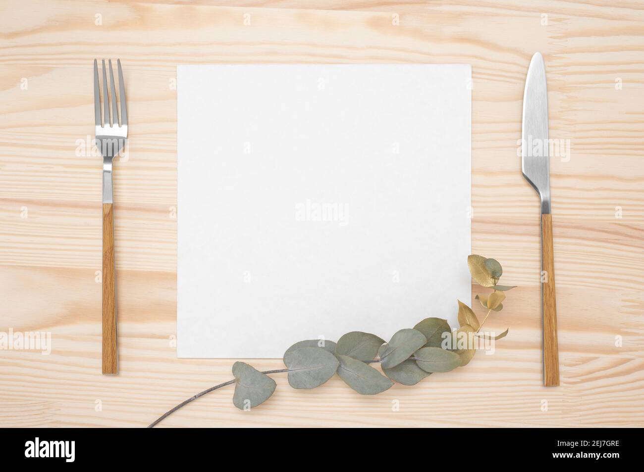 Coltello e forchetta con carta bianca per il menù o il testo della ricetta e branca di eucalipto d'oro su tavola di legno. Mockup carta bianca e posate su rustico tavolo di legno Foto Stock