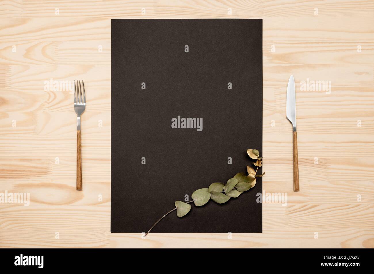 Coltello e forchetta con carta nera a4, a3 per testo di menu o ricette e rametto di eucalipto d'oro su tavola di legno. Mockup carta nera vuota e posate su rustico Foto Stock