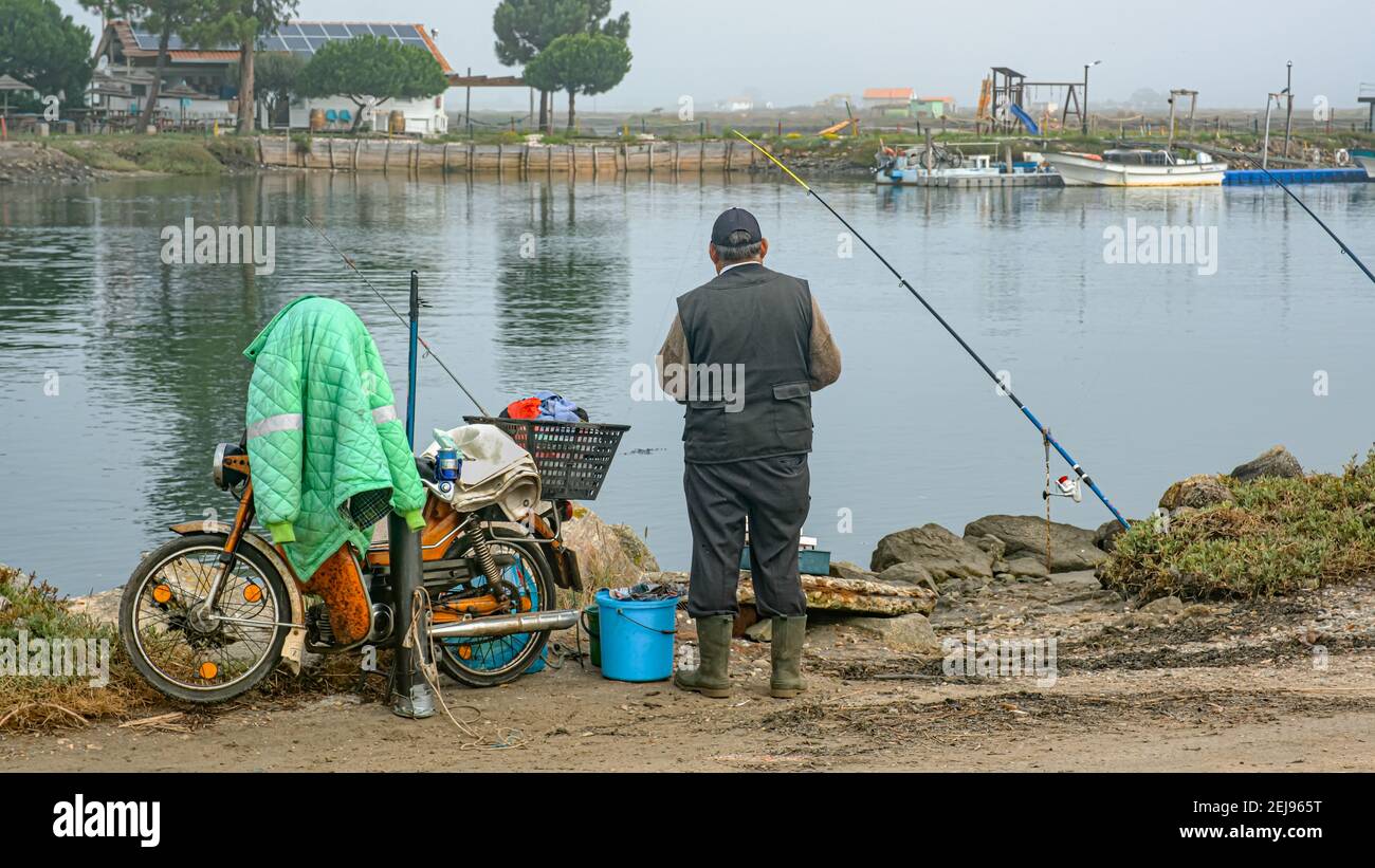 Uomo pesca con la cuffia sulla riva di un'insenatura con canne e attrezzatura, moto e giacca accanto a lui in Portogallo all'alba Foto Stock