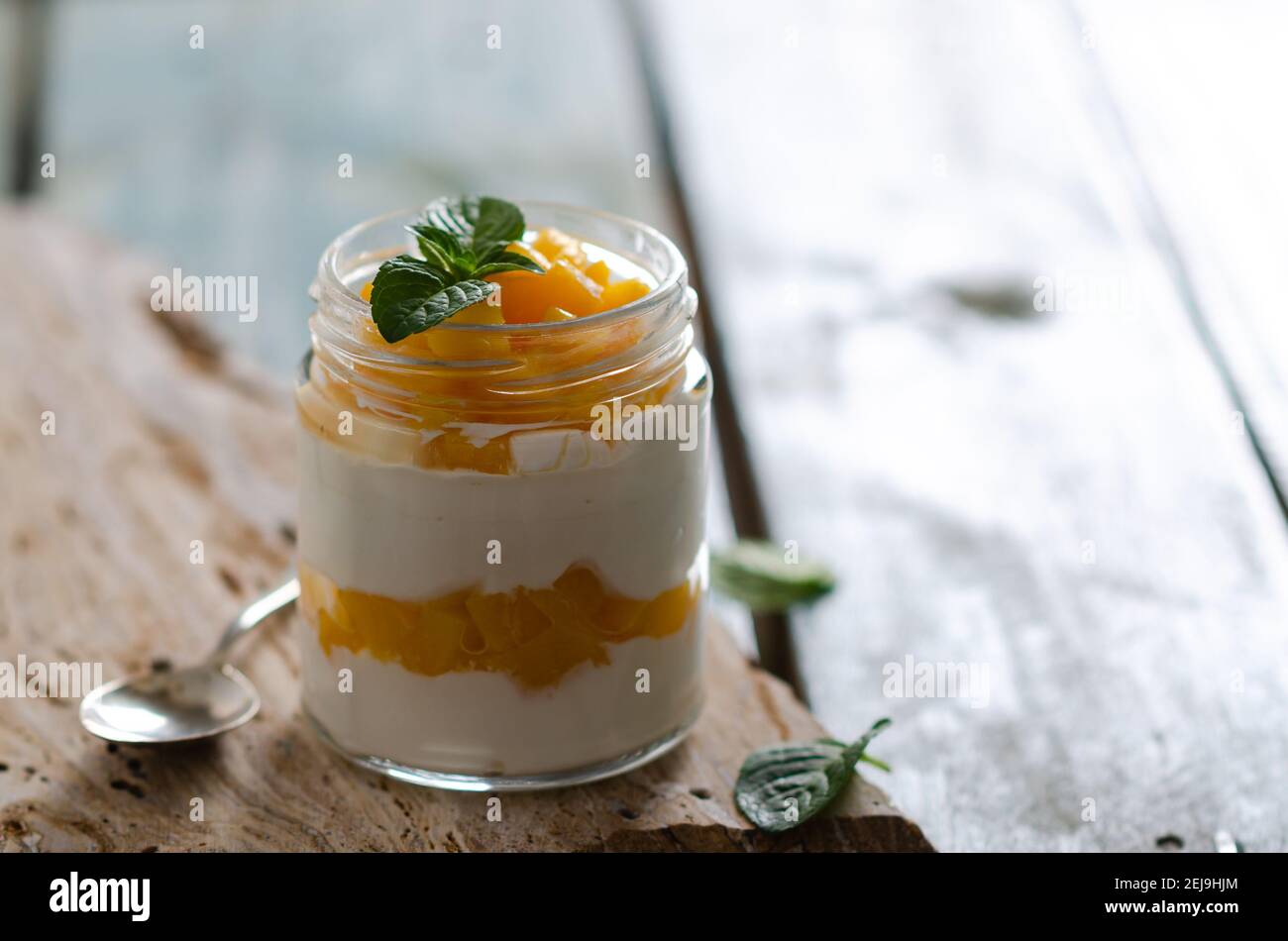 Yogurt bianco con pezzi di pesca e foglie di menta in un vasetto su una pietra gialla e sfondo di legno azzurro. Foto Stock