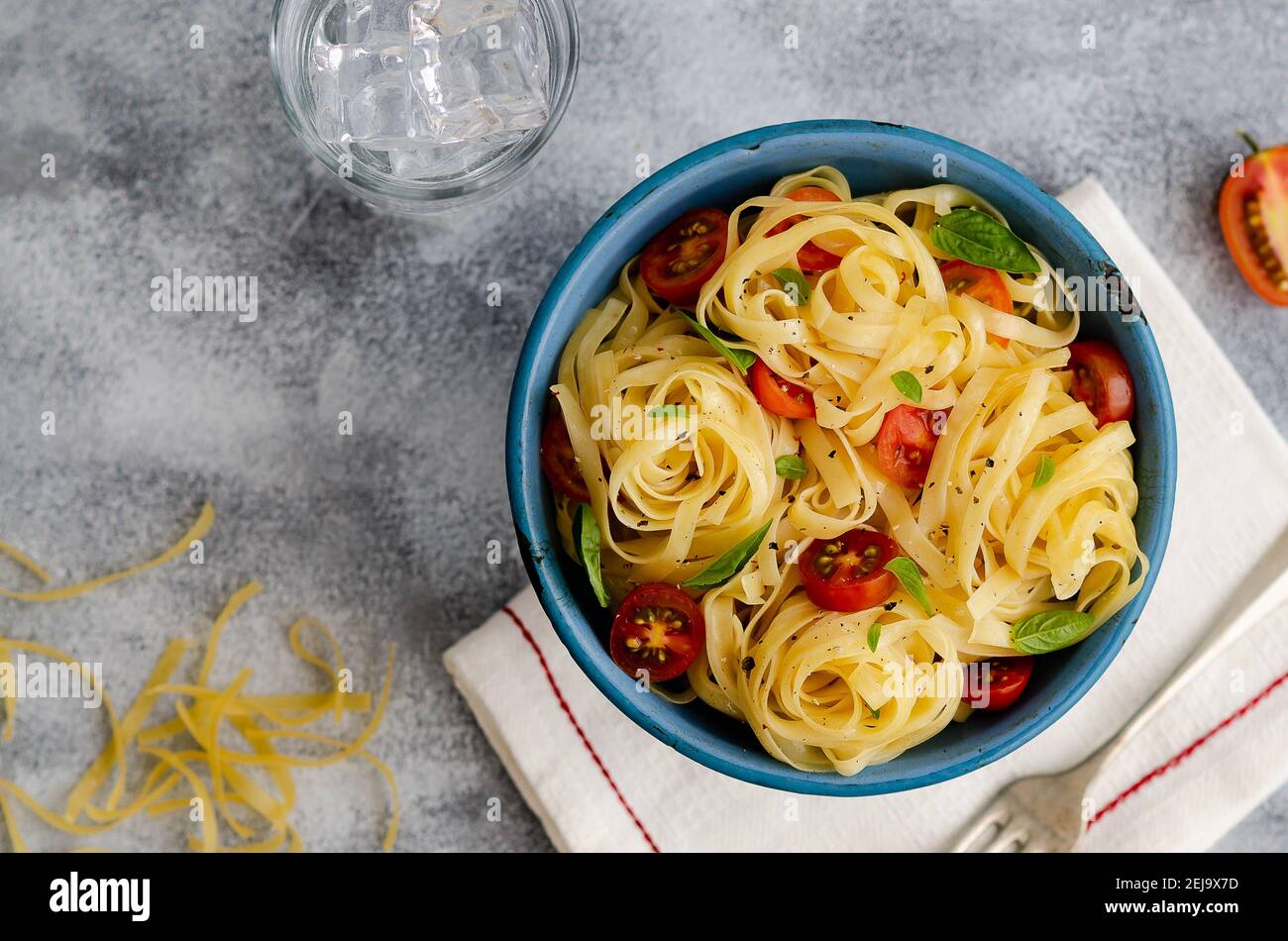 Fettuccine con pomodori ciliegini a fette e foglie di basilico in un piatto blu su sfondo grigio, con un tovagliolo bianco, forchetta, un bicchiere d'acqua e pasta cruda. Foto Stock