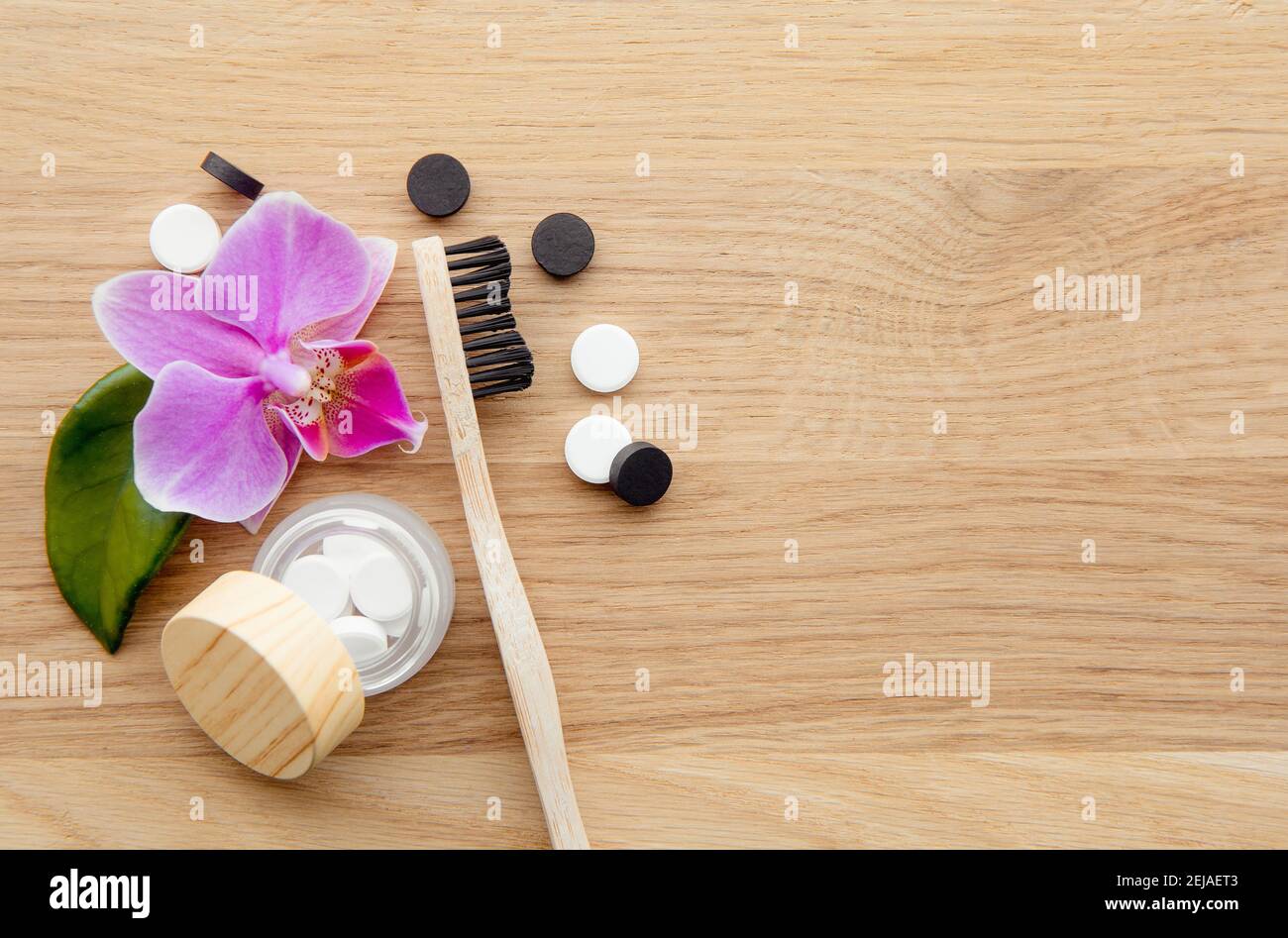 Vista dall'alto delle compresse di dentifricio al carbone attivo nero e al fluoruro bianco. Concetto di cura dentale. Sfondo minimalista in legno con fiori di orchidea rosa. Foto Stock