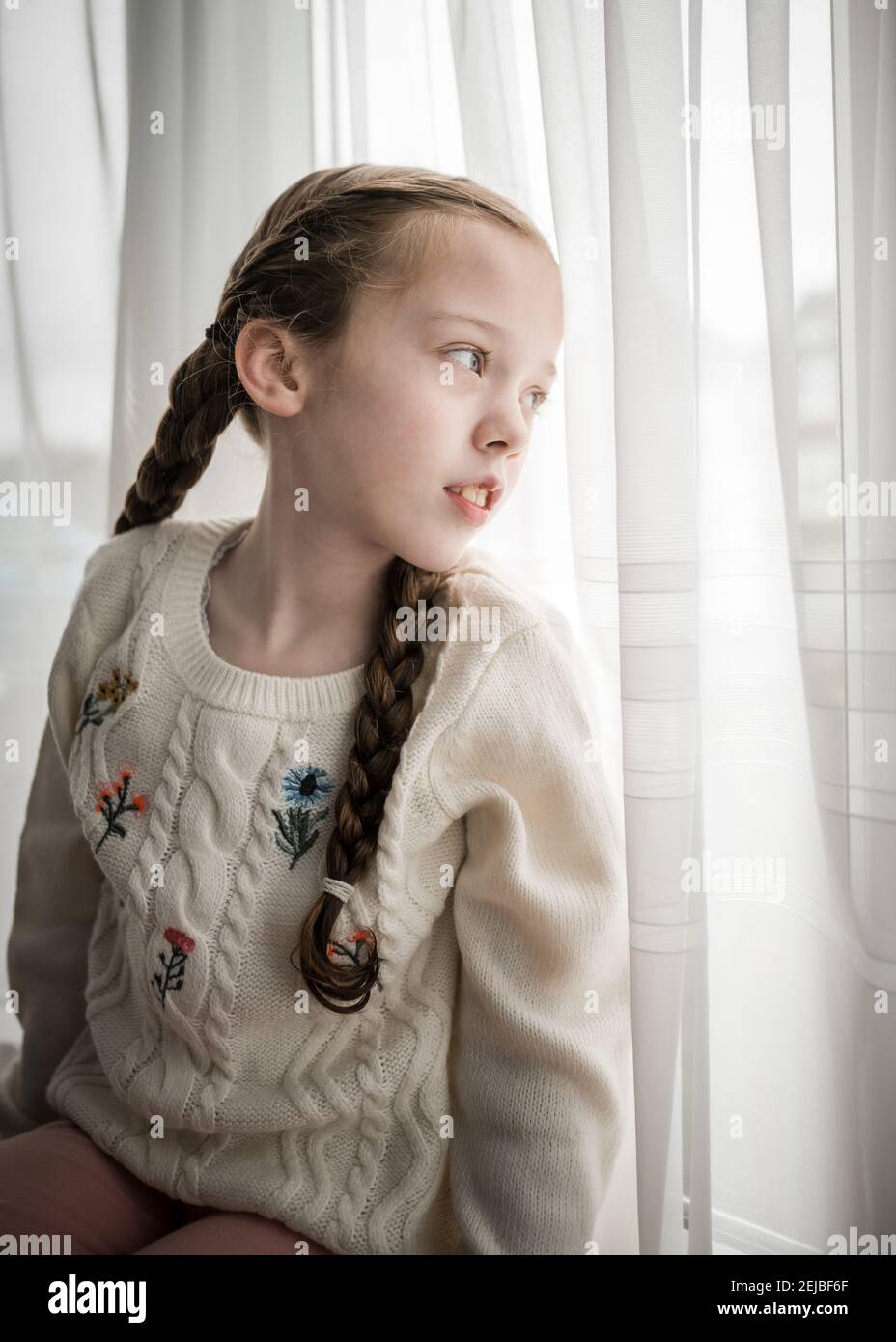 Giovane bella ragazza bloccata all'interno sedette sul windowsill guardando attraverso tenda finestra esterna con cute pigtail capelli lunghi estate brillante pandemia di blocco Foto Stock