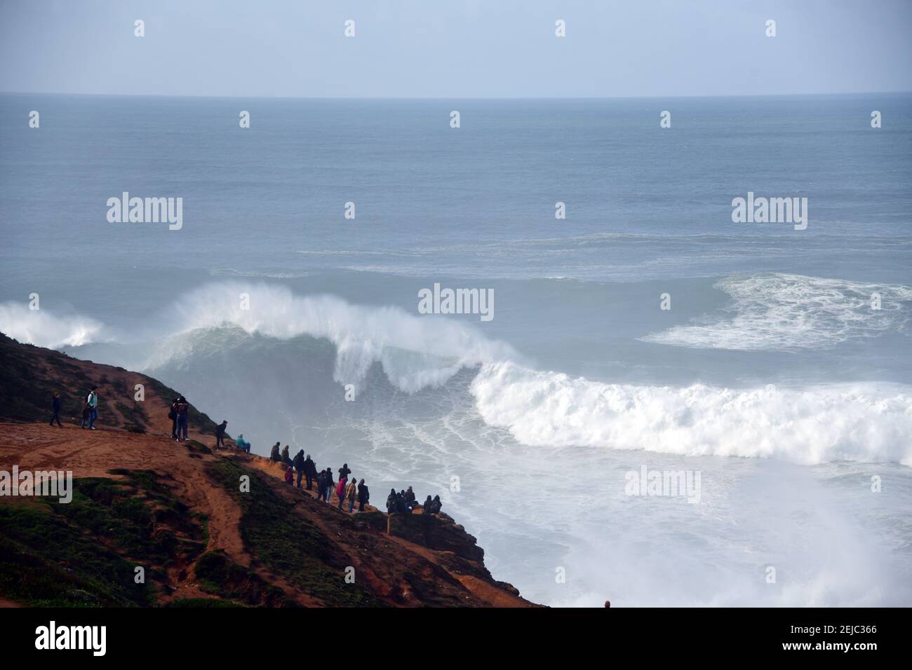 Piccola folla che guarda grandi onde dalle scogliere di Nazare Portogallo Foto Stock