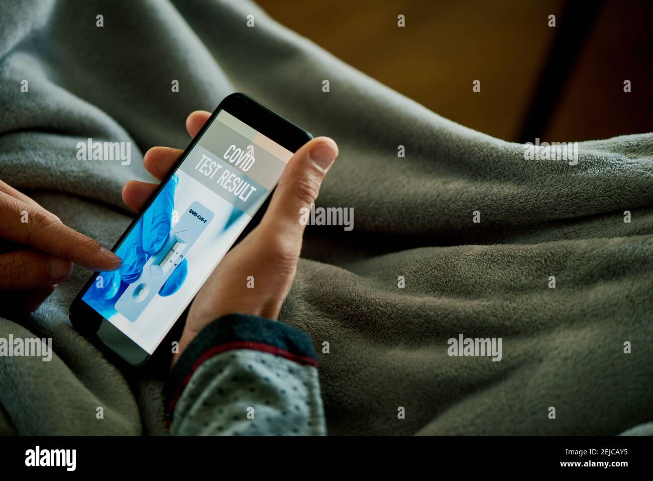 un giovane caucasico in pigiama, seduto su un divano avvolto in una coperta grigia, sta per verificare il risultato del suo covid test in un'app simulata in hi Foto Stock