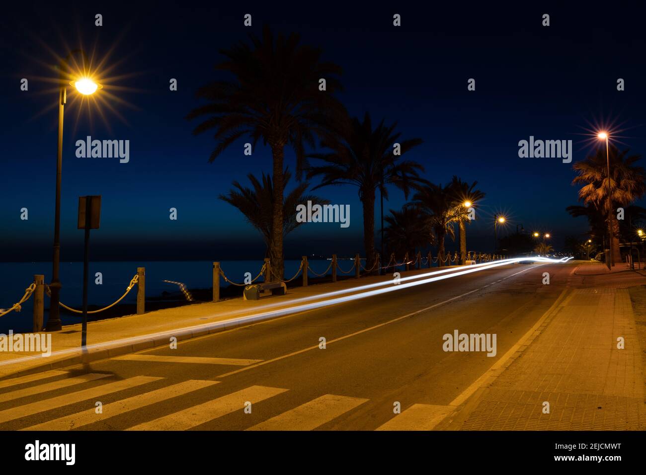 Una lunga esposizione vista notturna della strada fronte oceano con linee chiare da auto e palme in silhouette e l'oceano sullo sfondo Foto Stock