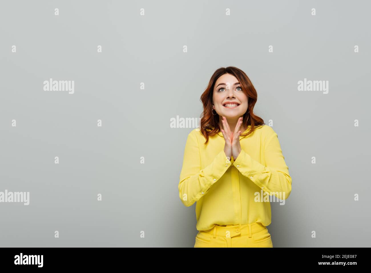 gioiosa donna in camicia gialla che guarda e si aggrappano le mani su grigio Foto Stock