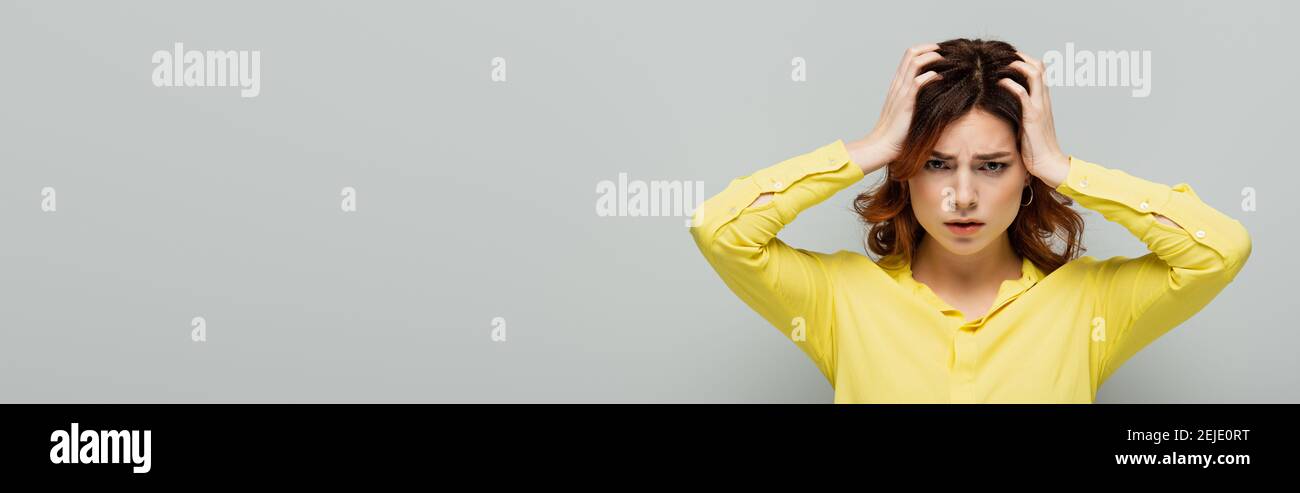 donna triste in camicia gialla che tocca la testa mentre guarda la macchina fotografica su grigio, banner Foto Stock
