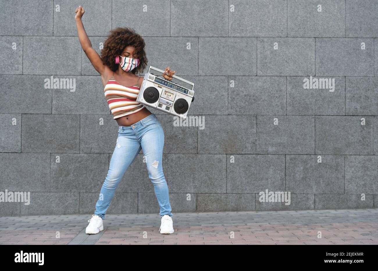 Giovane donna afro che indossa maschera danzante all'aperto mentre ascolti alla musica con le cuffie wireless e stereo stereo stereo stereo con stereo portatile vintage Foto Stock