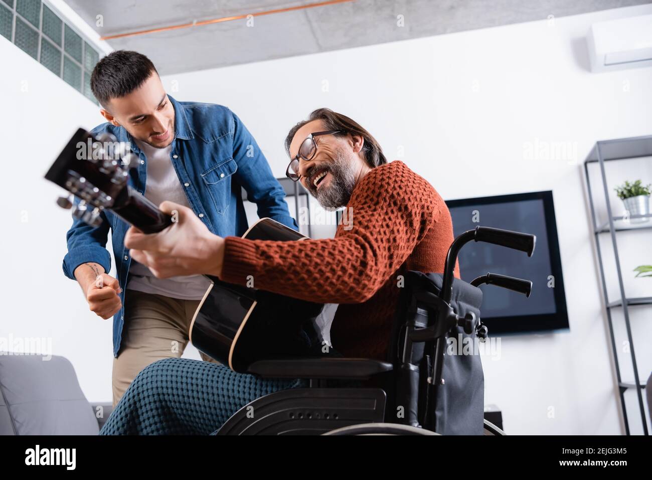 vista ad angolo basso di un uomo allegro e disabile che suona la chitarra vicino eccitato figlio ispanico Foto Stock