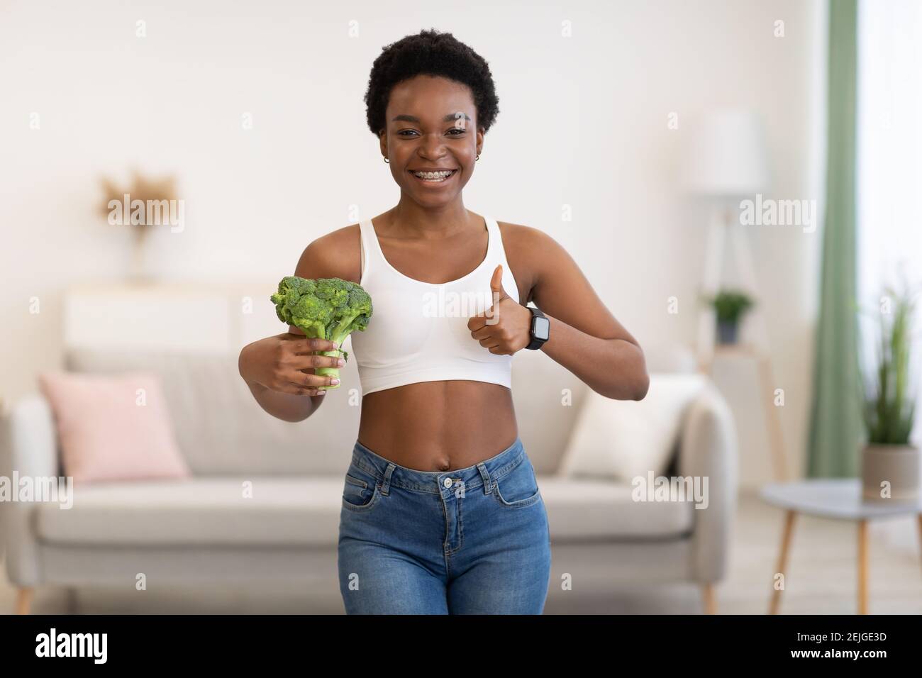 Happy Slim Black Woman gesturing Thumbs-Up Holding Broccoli a casa Foto Stock