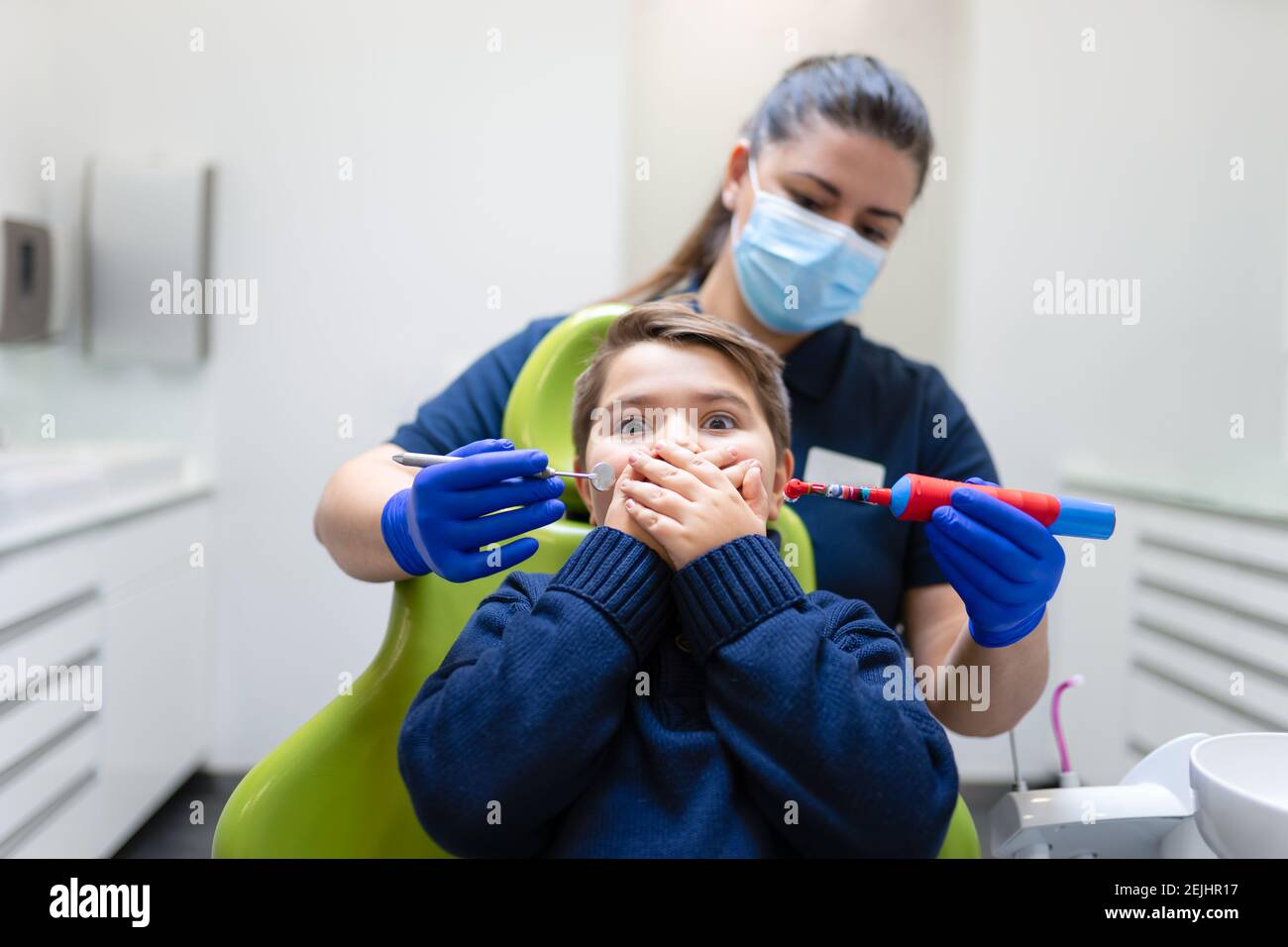 Il dentista tiene la spazzola elettrica e lo specchio vicino alla bocca del ragazzo. Teen non vuole fare igiene orale. Concetto di odontoiatria pediatrica Foto Stock