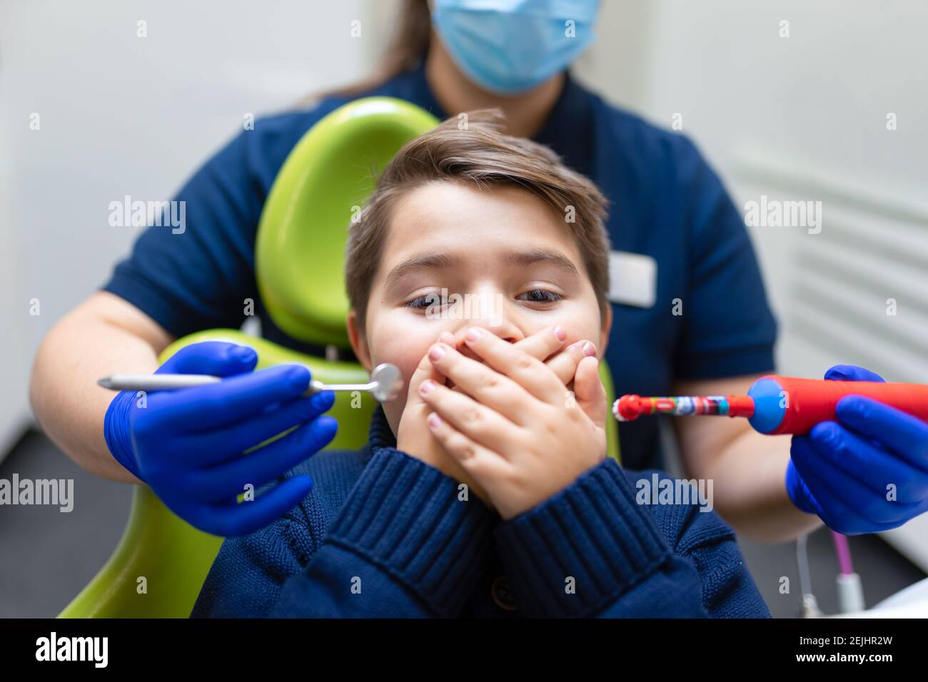 Il dentista tiene la spazzola elettrica e lo specchio vicino alla bocca del ragazzo. Teen non vuole fare igiene orale. Concetto di odontoiatria pediatrica Foto Stock