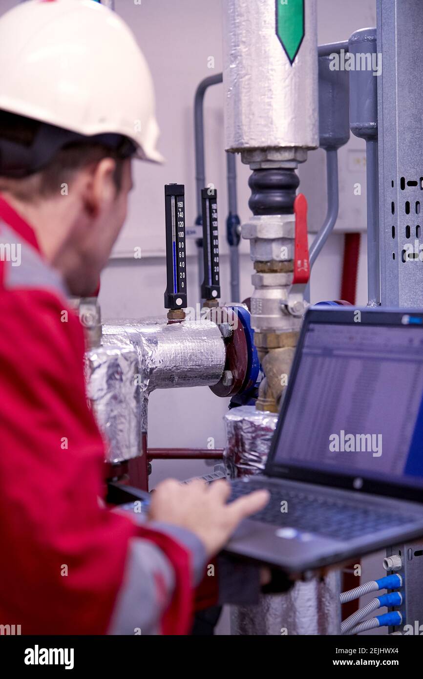 Tema del settore energetico: Un tecnico vestito di rosso e un casco bianco che controlla i parametri di riscaldamento su un computer portatile. Foto Stock