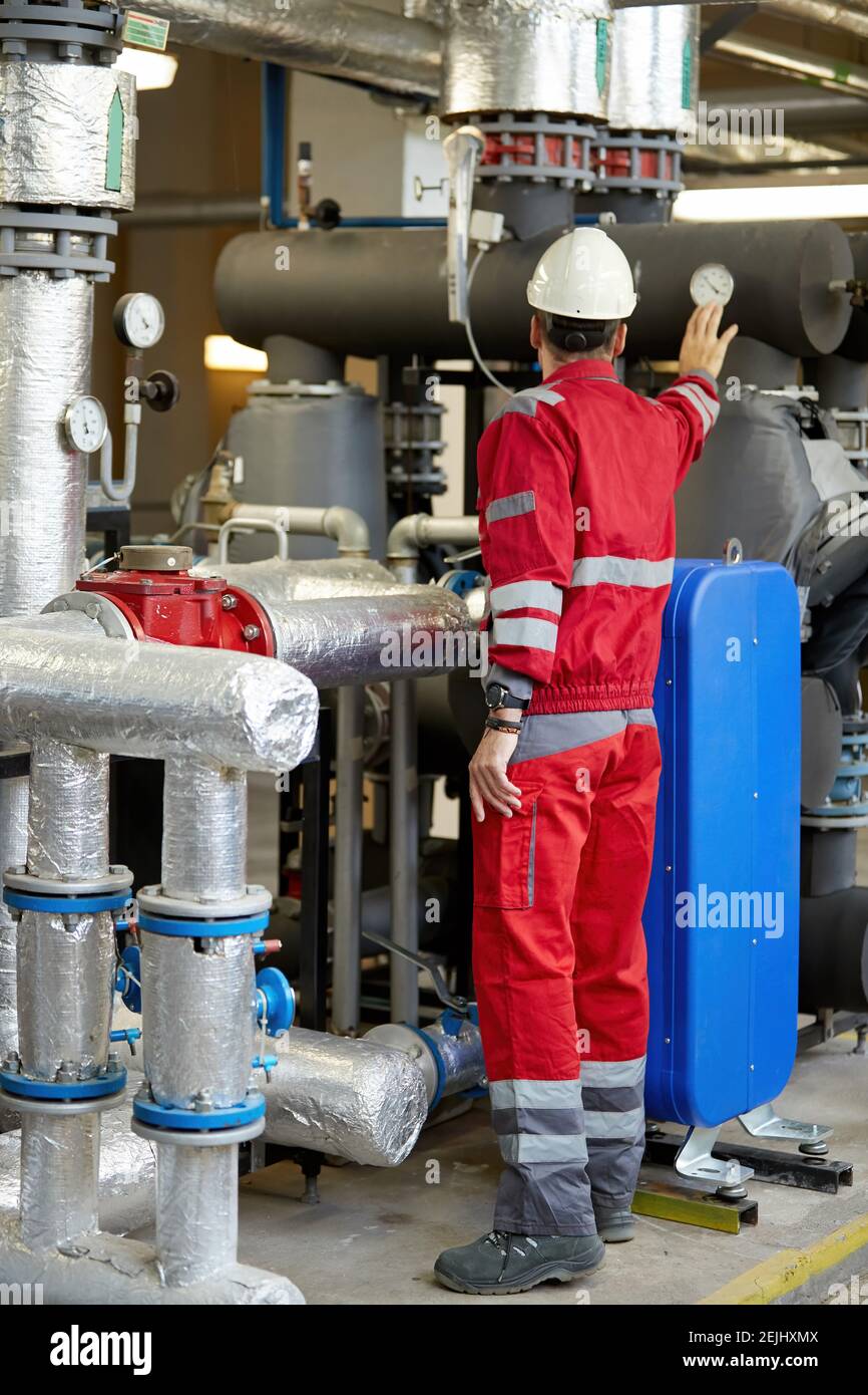 Tema del settore energetico: Un tecnico vestito con tute rosse e un casco bianco che controlla i parametri di riscaldamento. Tecnologia di distribuzione dell'acqua di riscaldamento. Foto Stock
