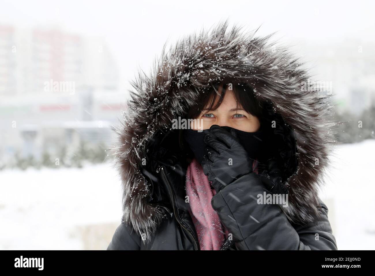 Tempo freddo, donna in maschera medica e cappuccio in pelliccia in piedi sulla strada invernale durante la neve. Concetto di gelo, malattia, febbre, protezione del coronavirus Foto Stock