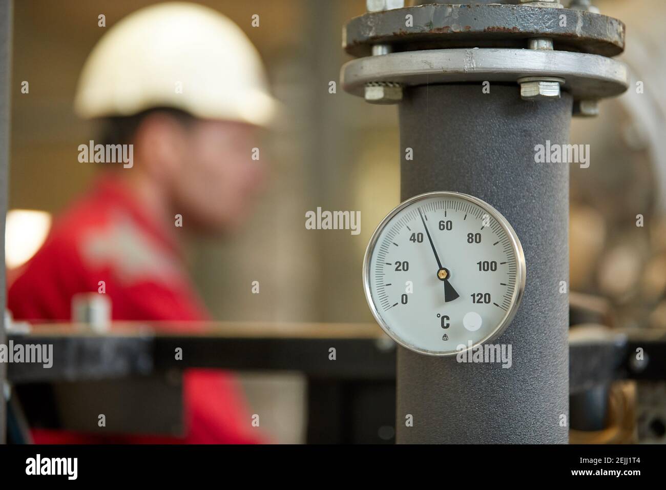 Termometro circolare sul tubo di alimentazione dell'acqua calda, impianto di riscaldamento. Tecnico che indossa una giacca rossa con un casco bianco sfocato sullo sfondo. Foto Stock
