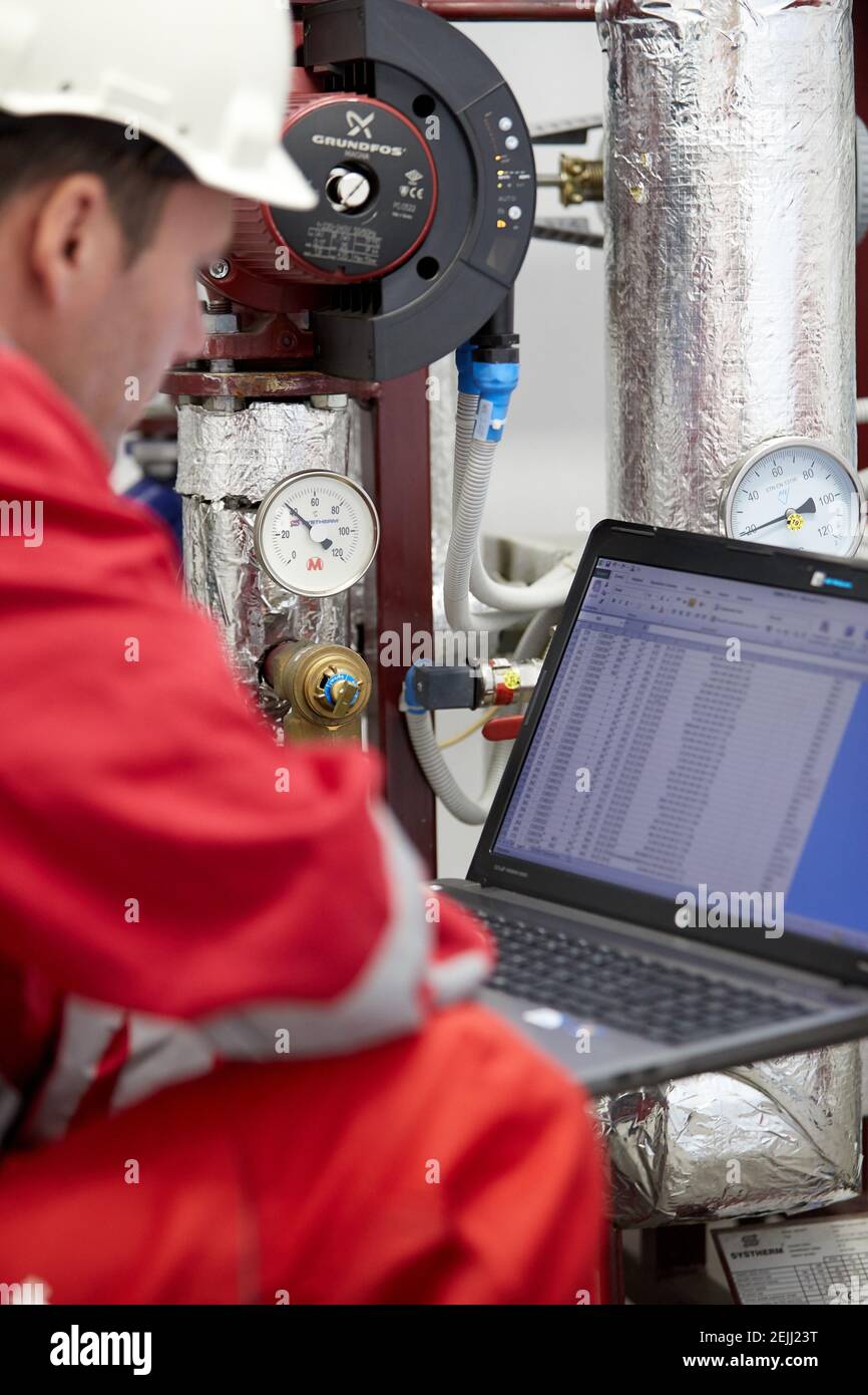 Tema del settore energetico: Un tecnico in rosso e un casco bianco controlla i parametri di riscaldamento su un computer portatile. Manutenzione e ispezione. Foto Stock