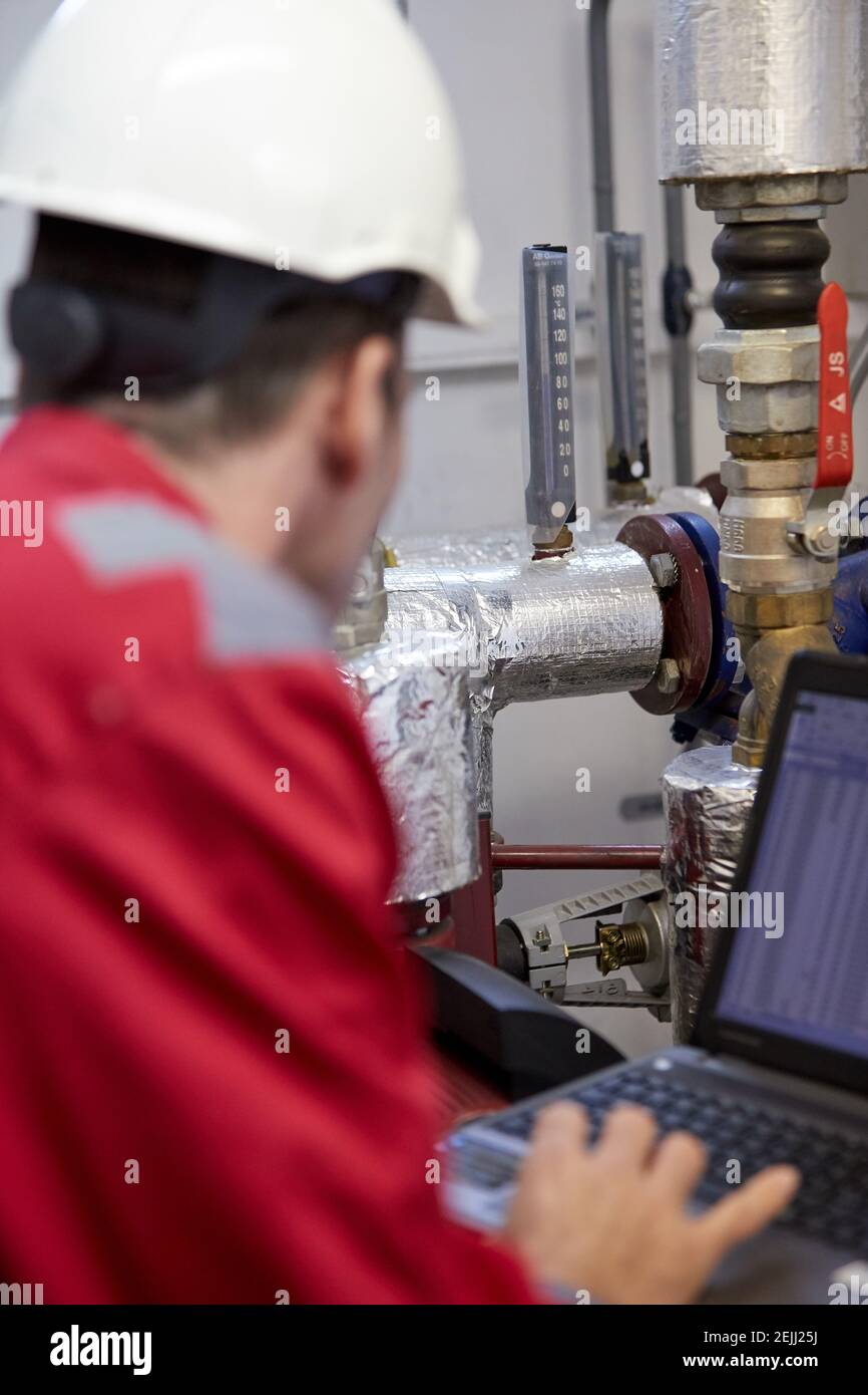 Tema del settore energetico: Un tecnico in rosso e un casco bianco controlla i parametri di riscaldamento su un computer portatile. Manutenzione e ispezione. Foto Stock