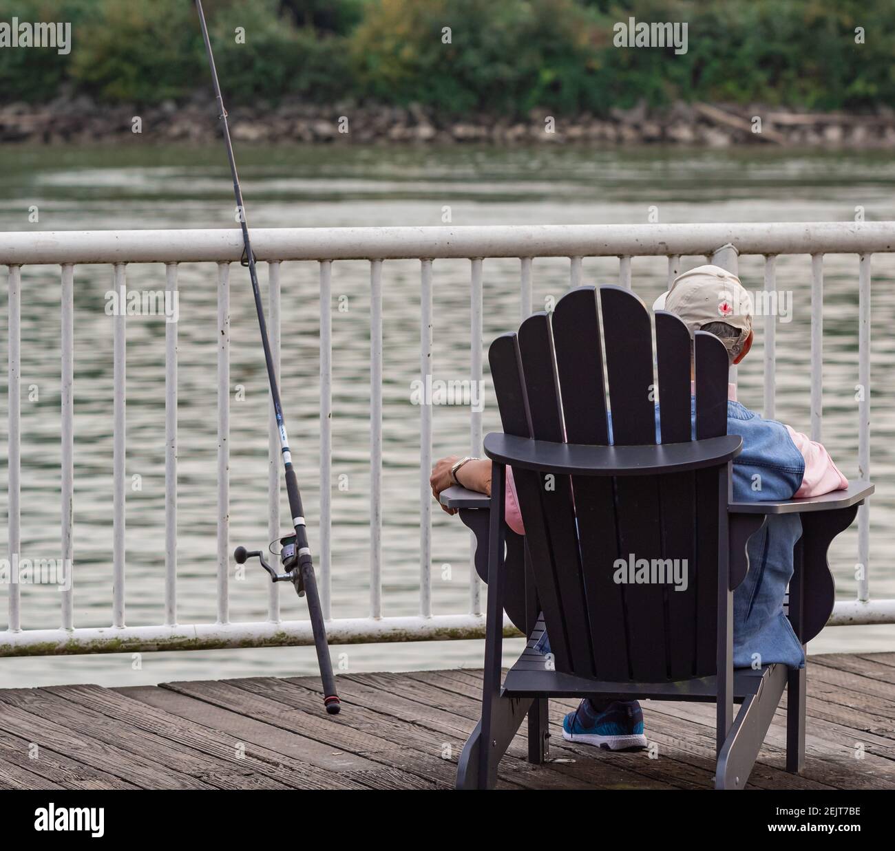 Vista posteriore di un vecchio uomo che pesca sul molo. Un anziano si siede su una sedia di legno vicino al fiume Fraser a New Westminster. Stile di vita attivo. Via Foto Stock